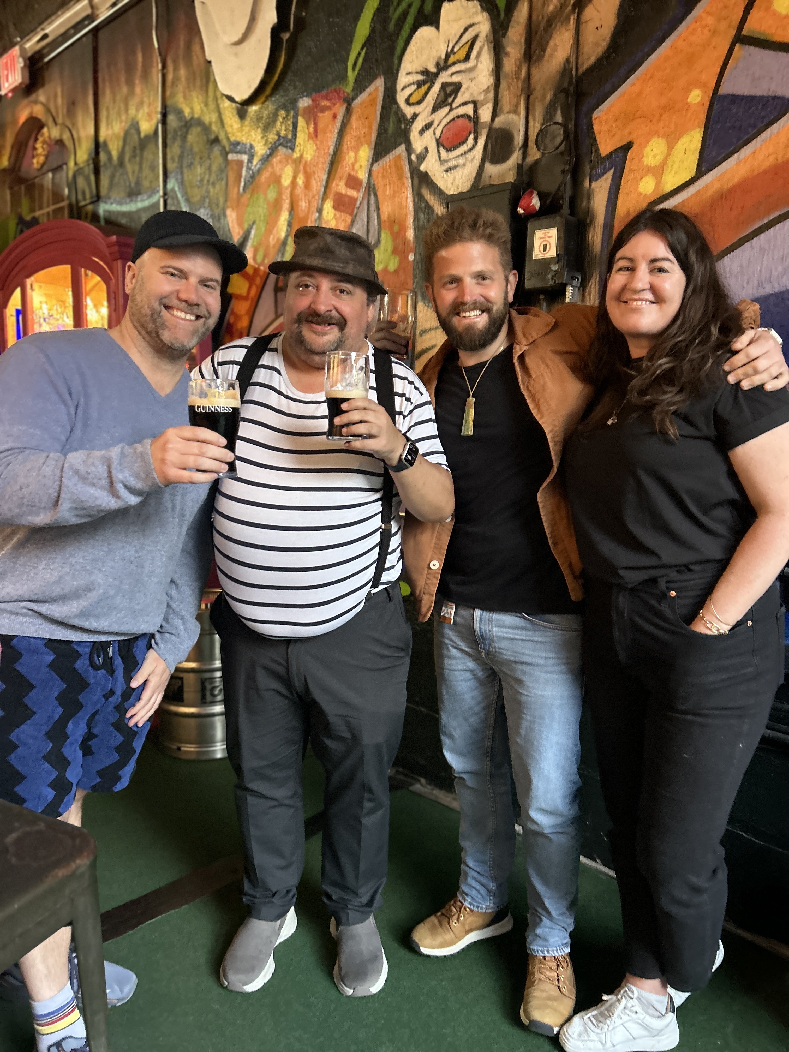 Four people stand closely together, smiling. Two hold dark beer glasses. They're in front of a colorful graffiti mural with vibrant, abstract designs.