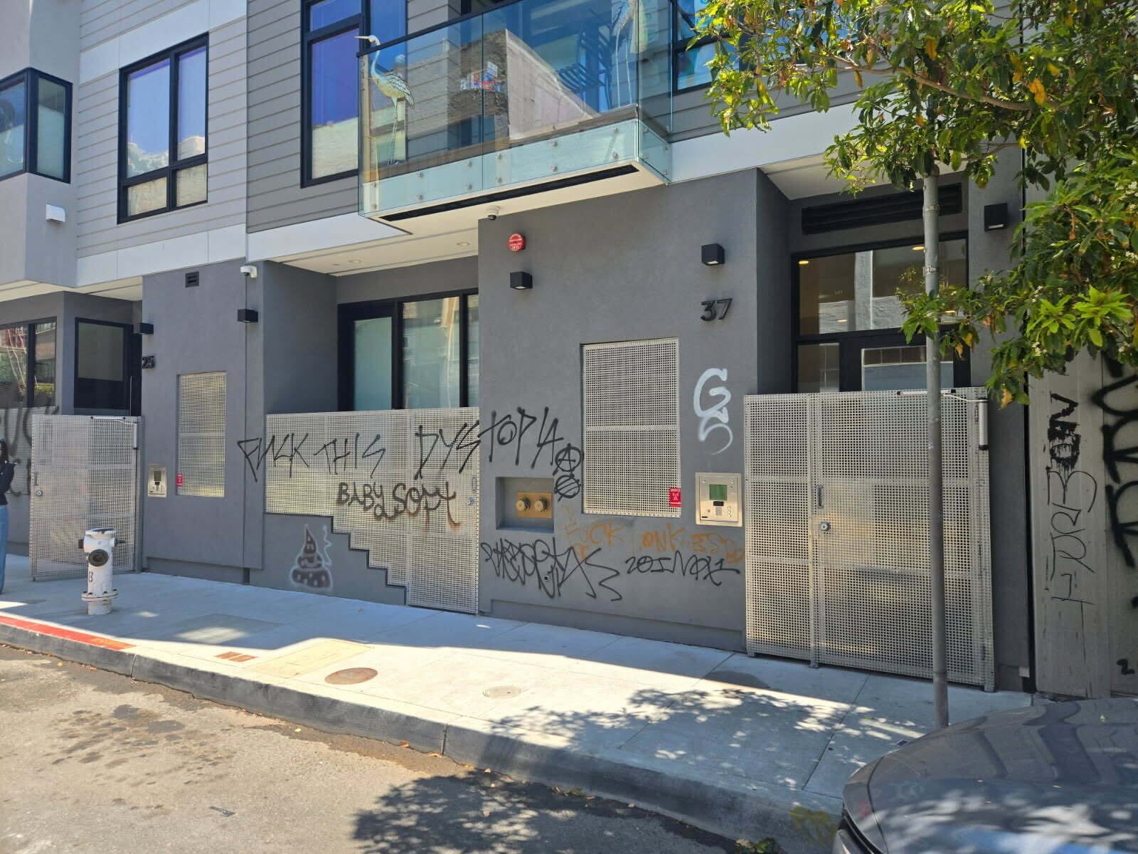 A modern urban building exterior with large windows is visible. There are metal gates and graffiti on the walls. A tree and a fire hydrant are in the foreground.