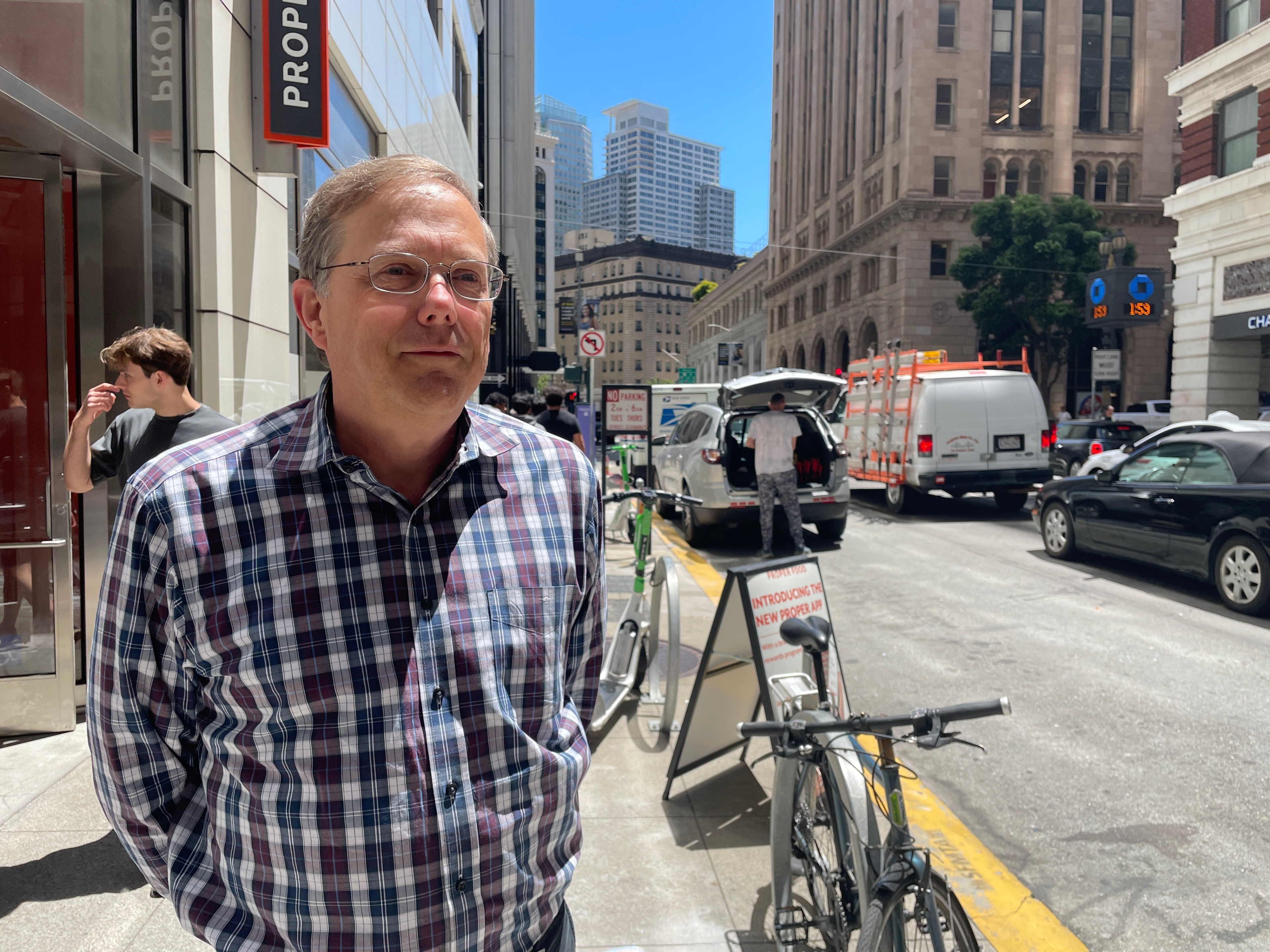 A man in a plaid shirt stands on a bustling city street with several vehicles and bicycles nearby. Tall buildings and a bright blue sky are in the background.