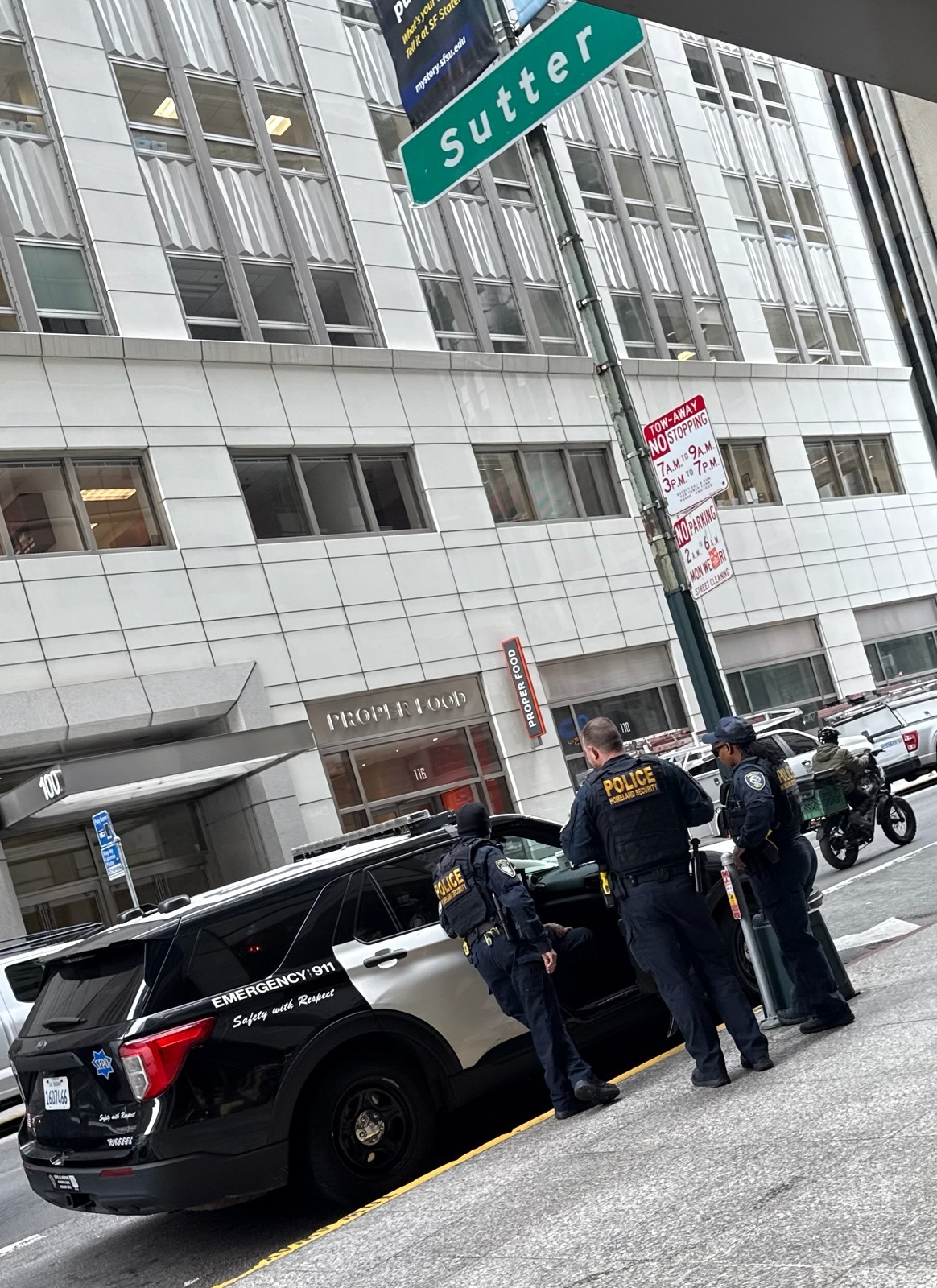 Three police officers stand beside a black and white police SUV on a city street, while a motorcyclist passes by. The street sign reads &quot;Sutter.&quot;