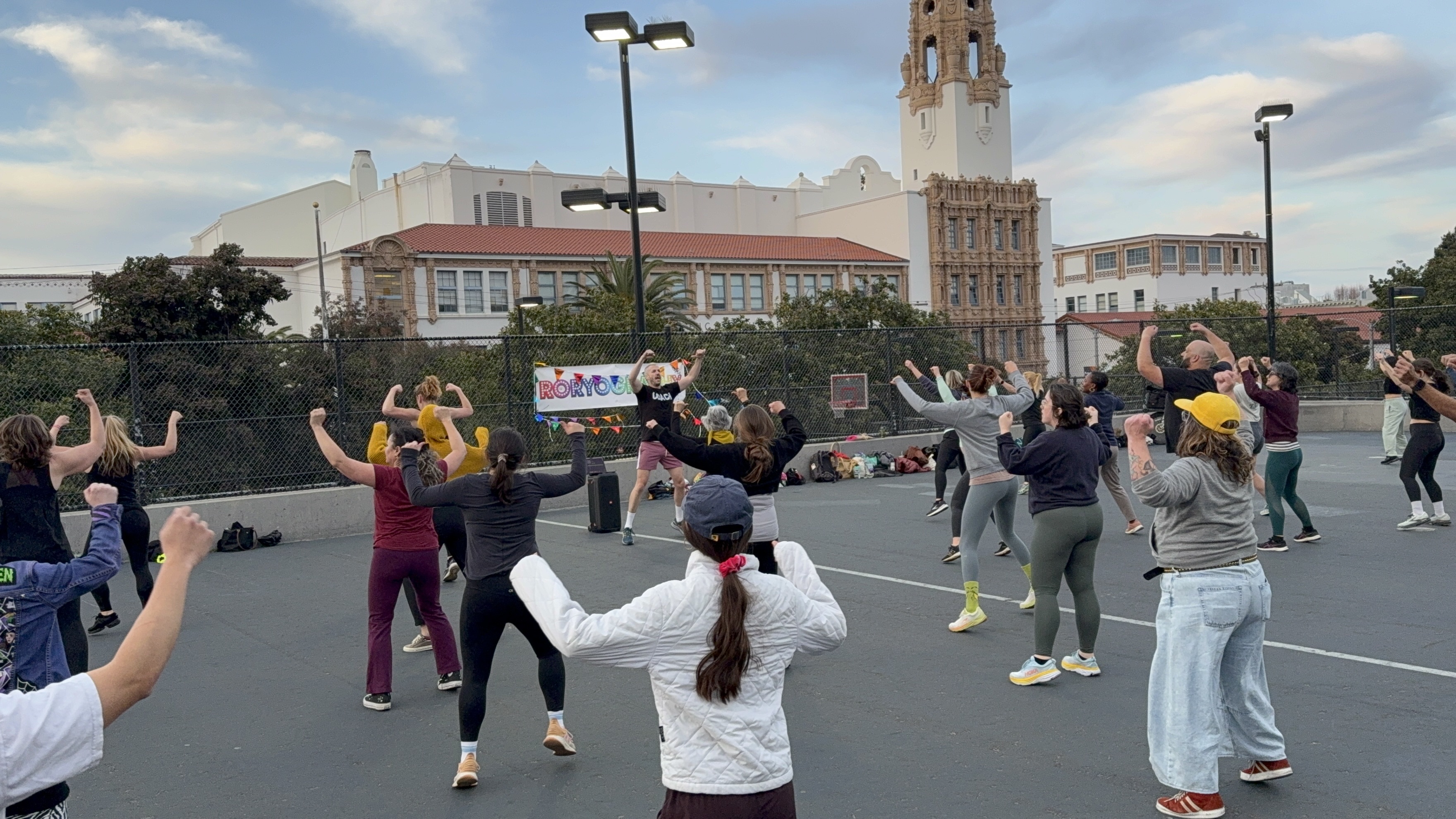 A group of people is engaged in an outdoor exercise class on a court, with a tall ornate building and a &quot;RORYOLOGY&quot; banner in the background.