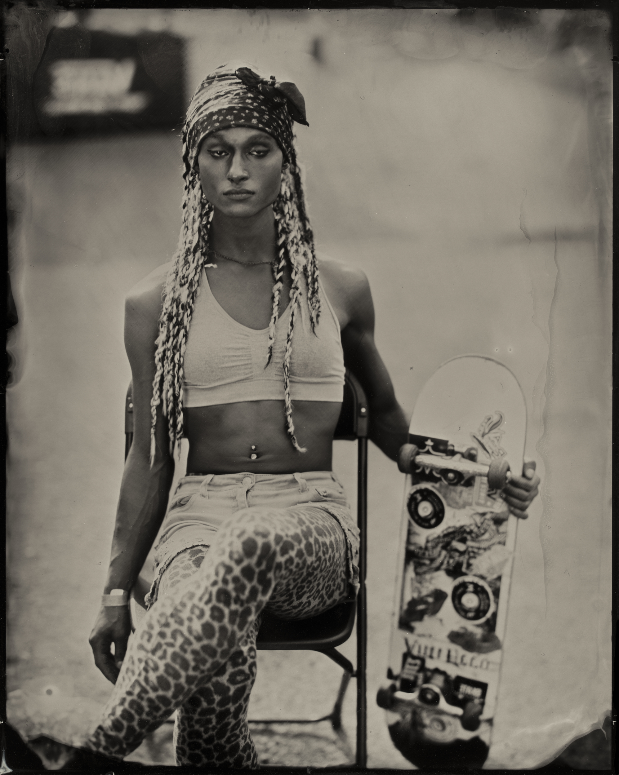 A person with braided hair and a headscarf sits on a chair with legs crossed, holding a skateboard. They're wearing a crop top, leopard print leggings, and shorts.