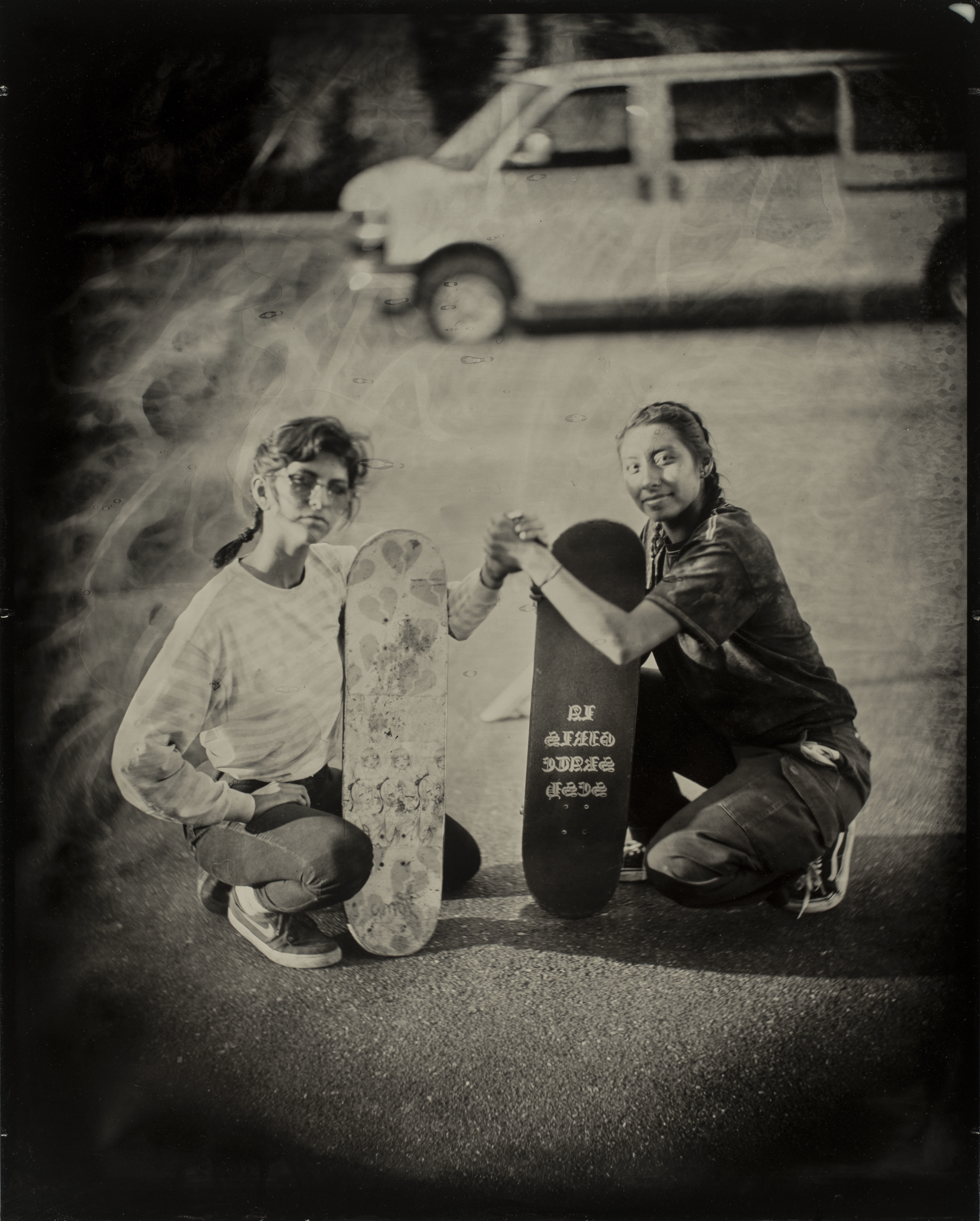 Two young people kneel on pavement, each holding a skateboard. They clasp hands, smiling, with a van in the blurred background. The photo is in a vintage style.