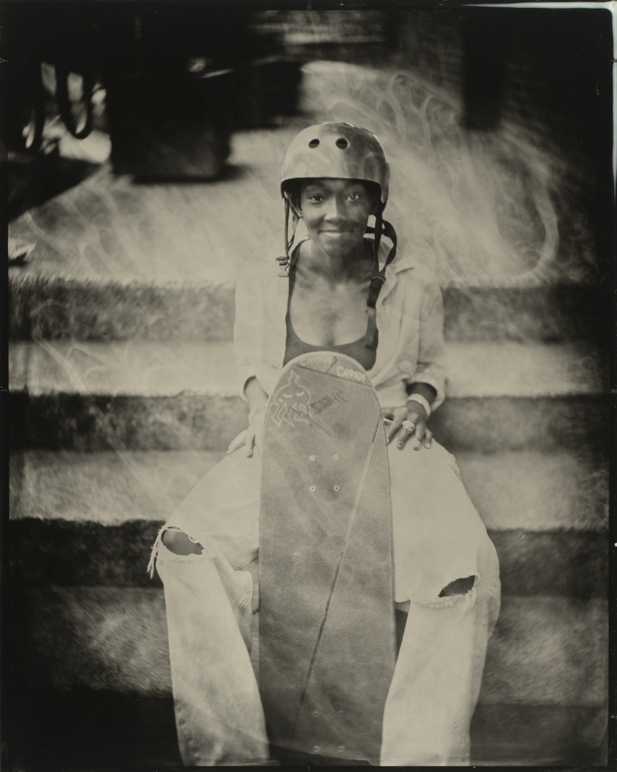 A person sits on steps wearing a helmet and holding a skateboard vertically. They have a faint smile and wear a jacket with ripped jeans.