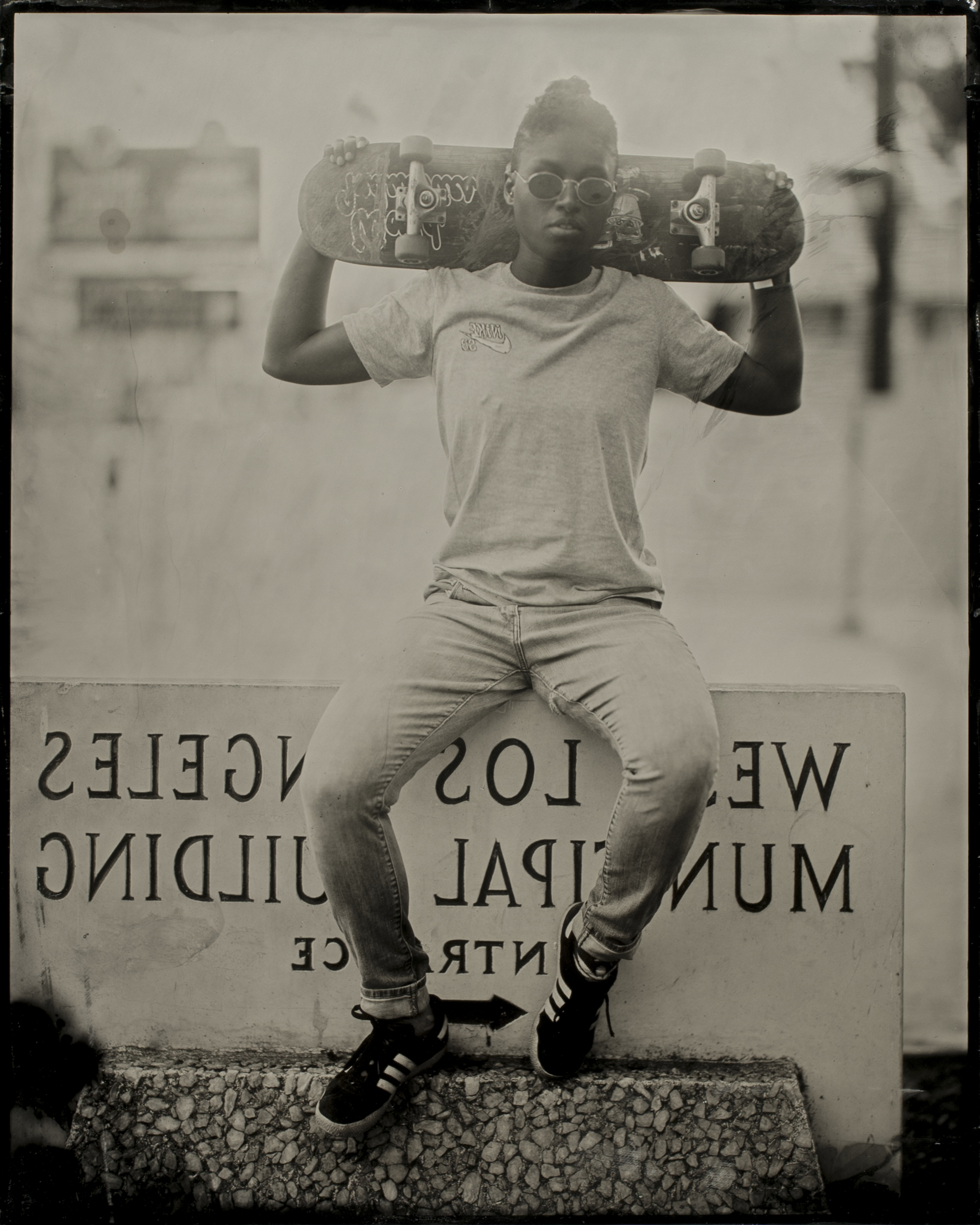 A person sits on a ledge holding a skateboard behind their neck. They're wearing sunglasses, a t-shirt, jeans, and sneakers. The background includes a reversed sign.