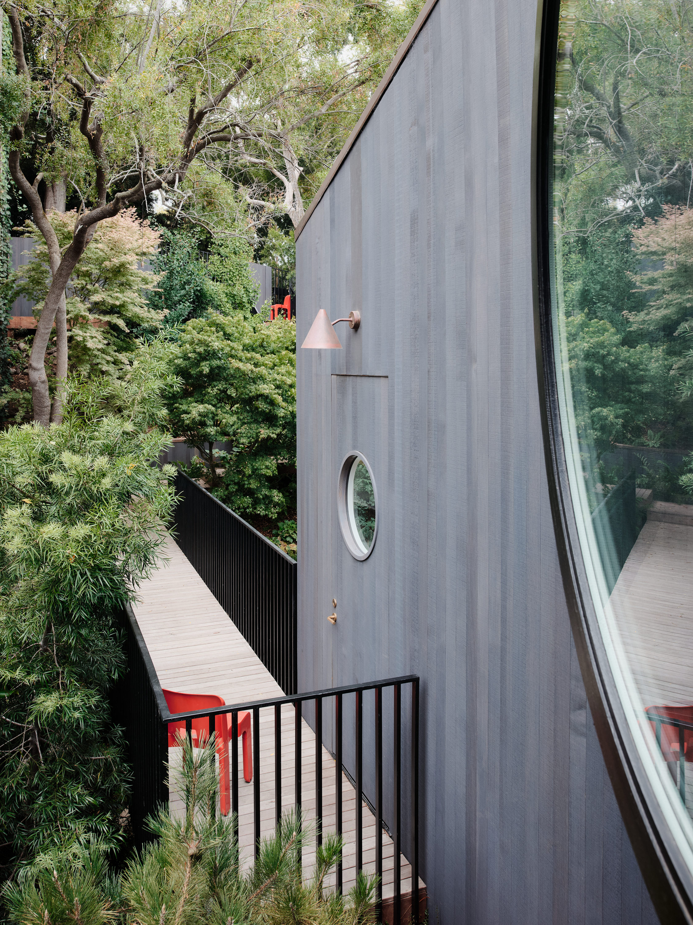A sleek, modern grey building with a large curved window and a round porthole window. A narrow wooden walkway with red chairs and surrounded by lush greenery.