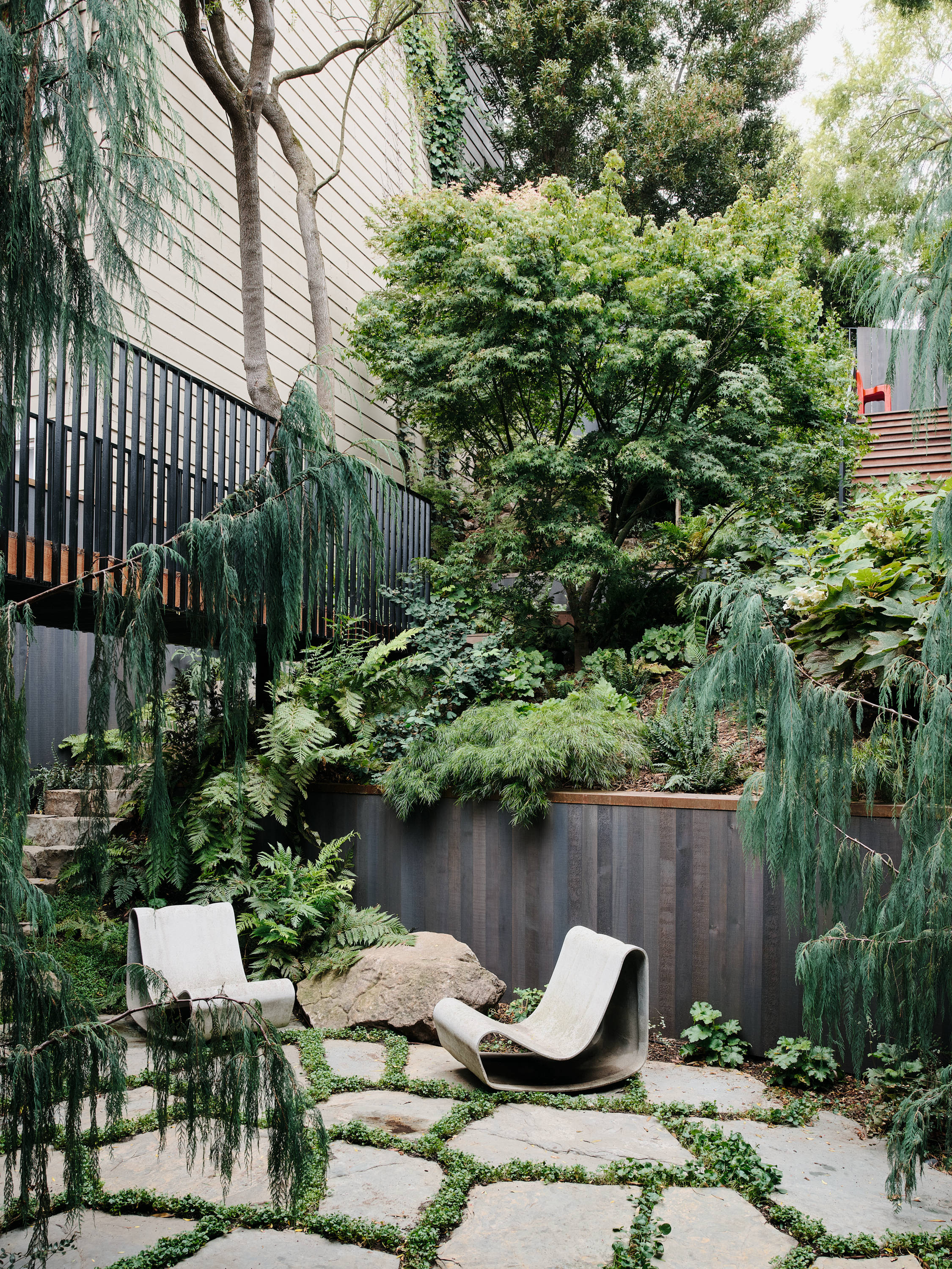 A serene garden with lush greenery, stone paths, two sculptural chairs, and wooden fencing. Tall trees and climbing plants add a natural, tranquil vibe.