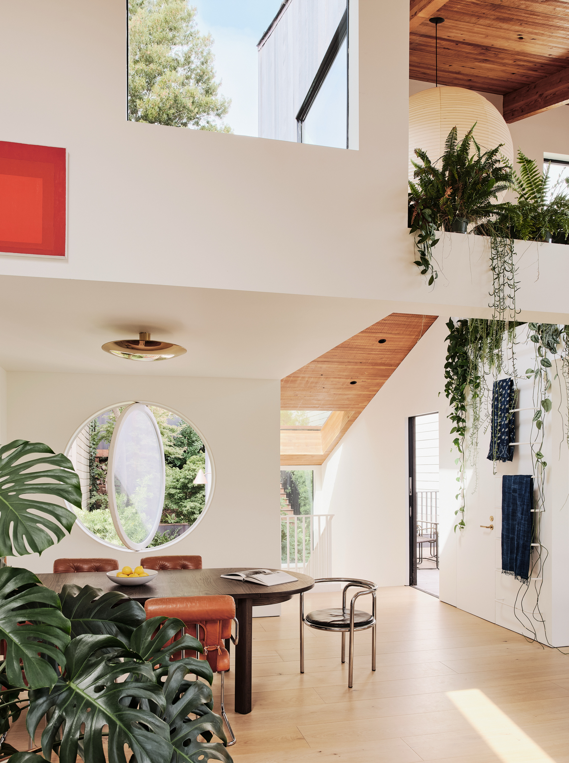 A modern dining area with a round table, leather chairs, and lush plants. Large windows and a skylight bring in natural light. A red painting adorns the wall.