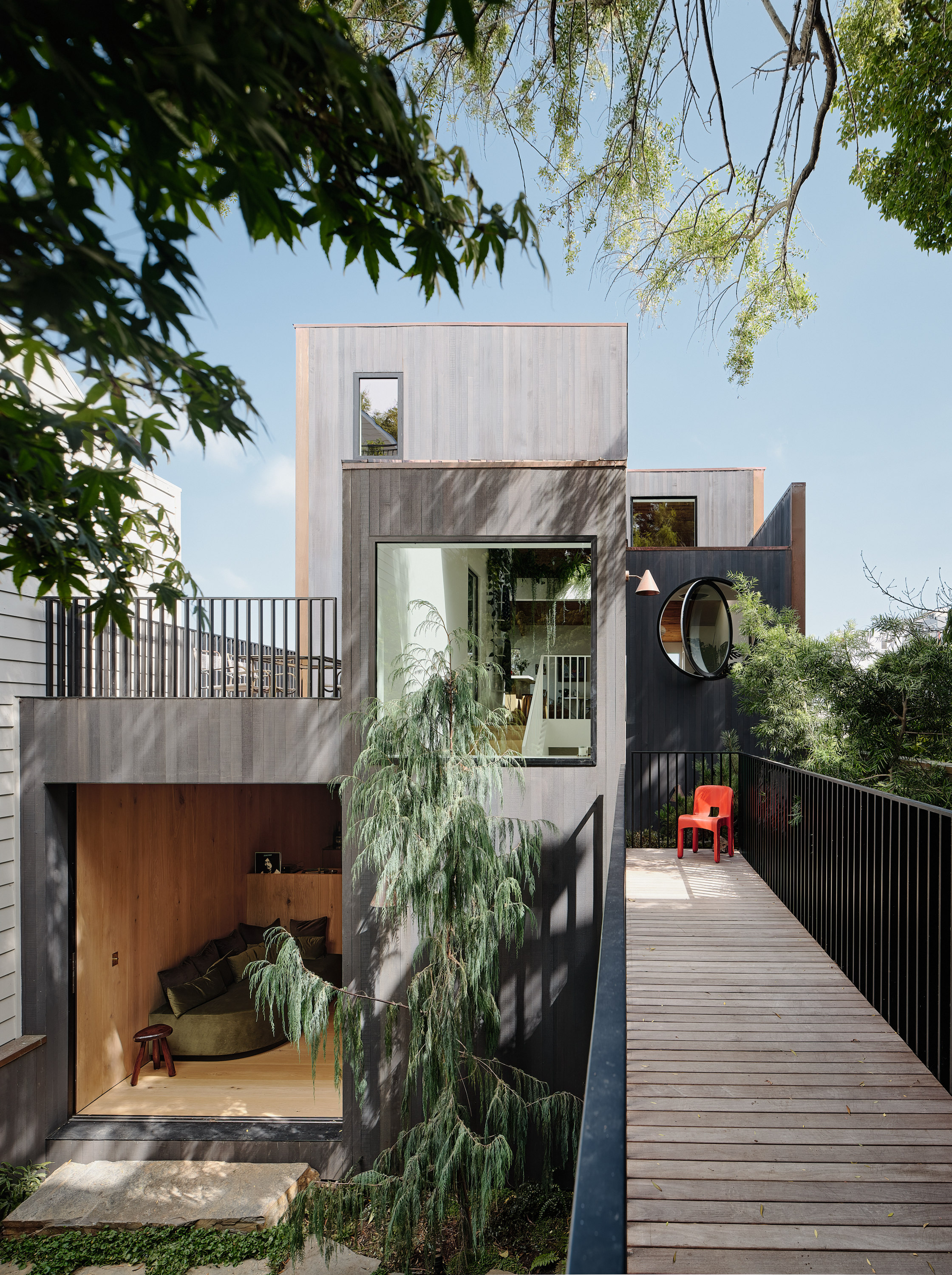 The image shows a modern, multi-level house with a wooden facade, large windows, and a wooden deck with a red chair. Trees and greenery surround the scene.