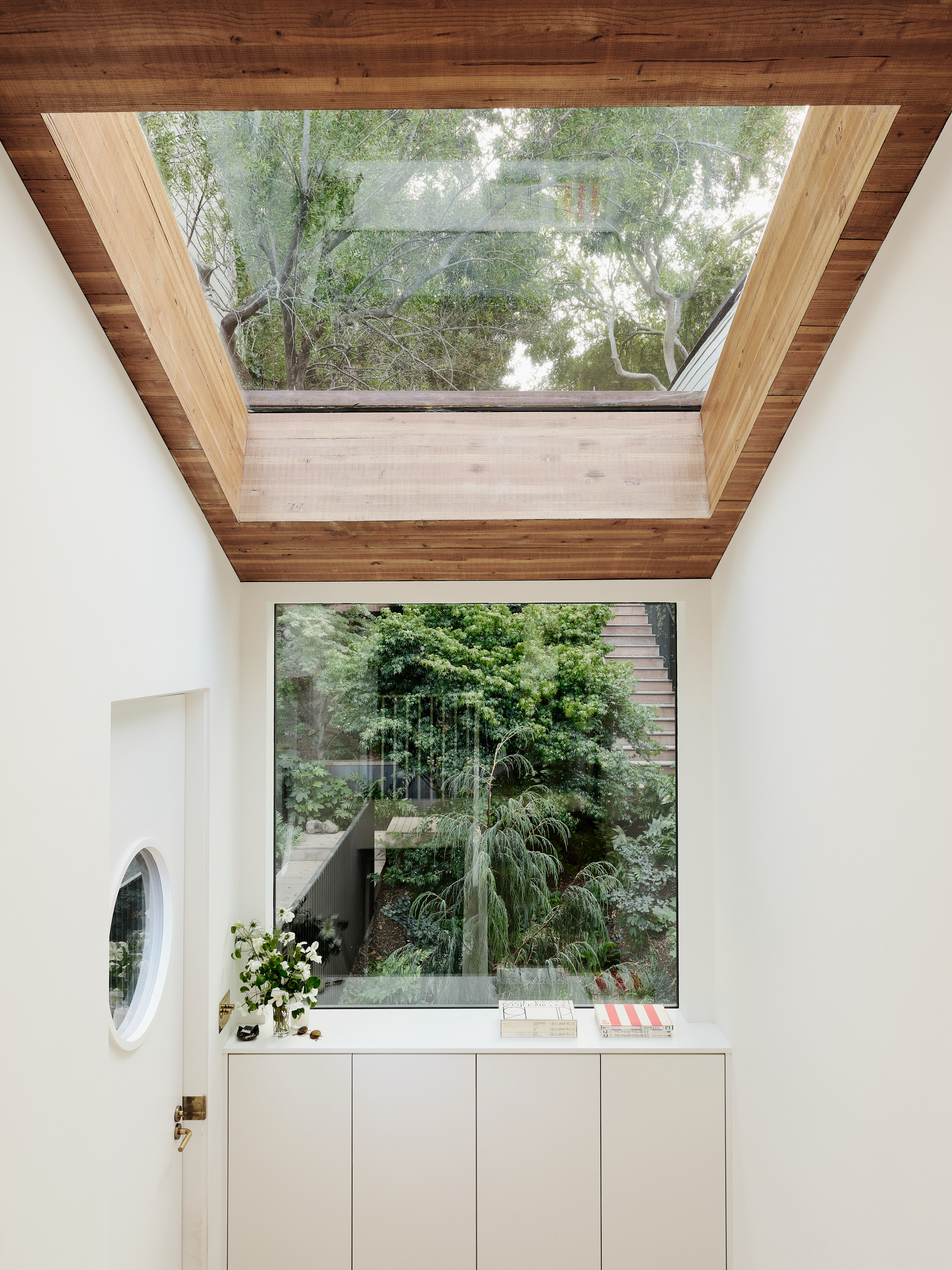 This image shows a hallway with a wooden ceiling and skylight, a large window overlooking lush greenery, and a white cabinet with books and flowers.
