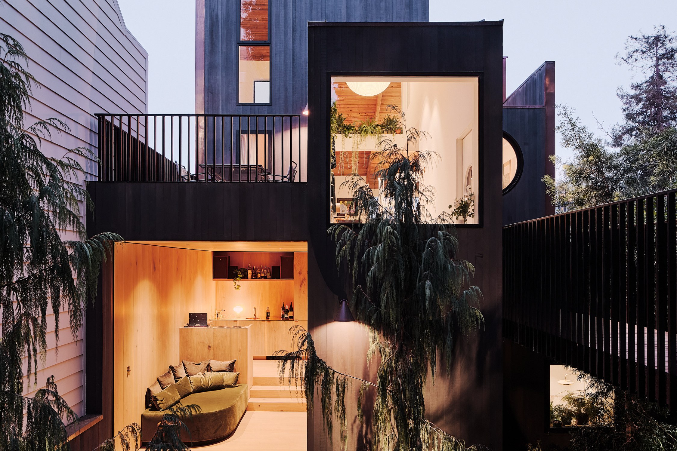 A modern, two-story house with large windows is surrounded by trees. The interior shows a cozy living area and a warm, softly lit kitchen.
