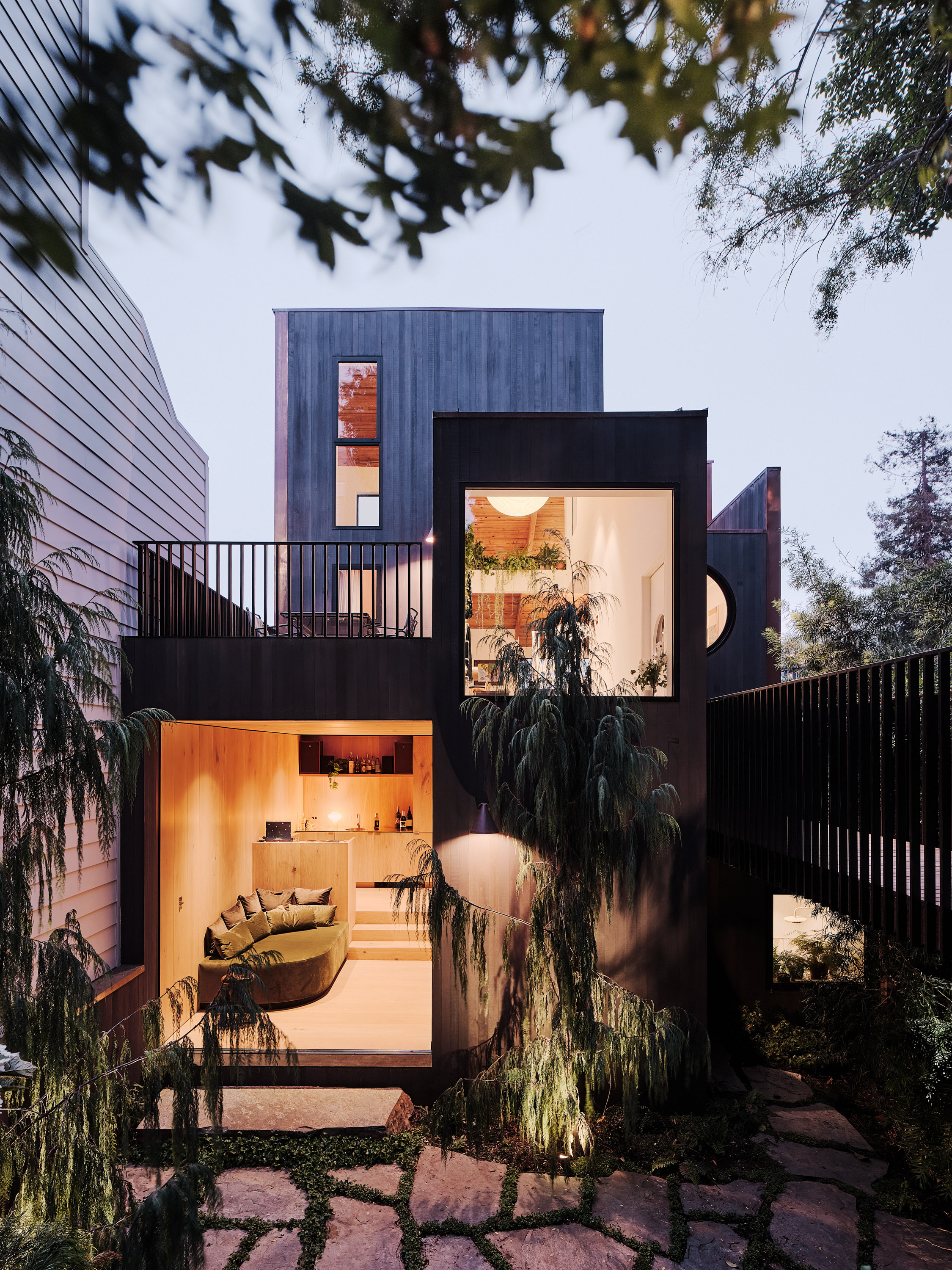 A modern, two-story house with large windows is surrounded by trees. The interior shows a cozy living area and a warm, softly lit kitchen.