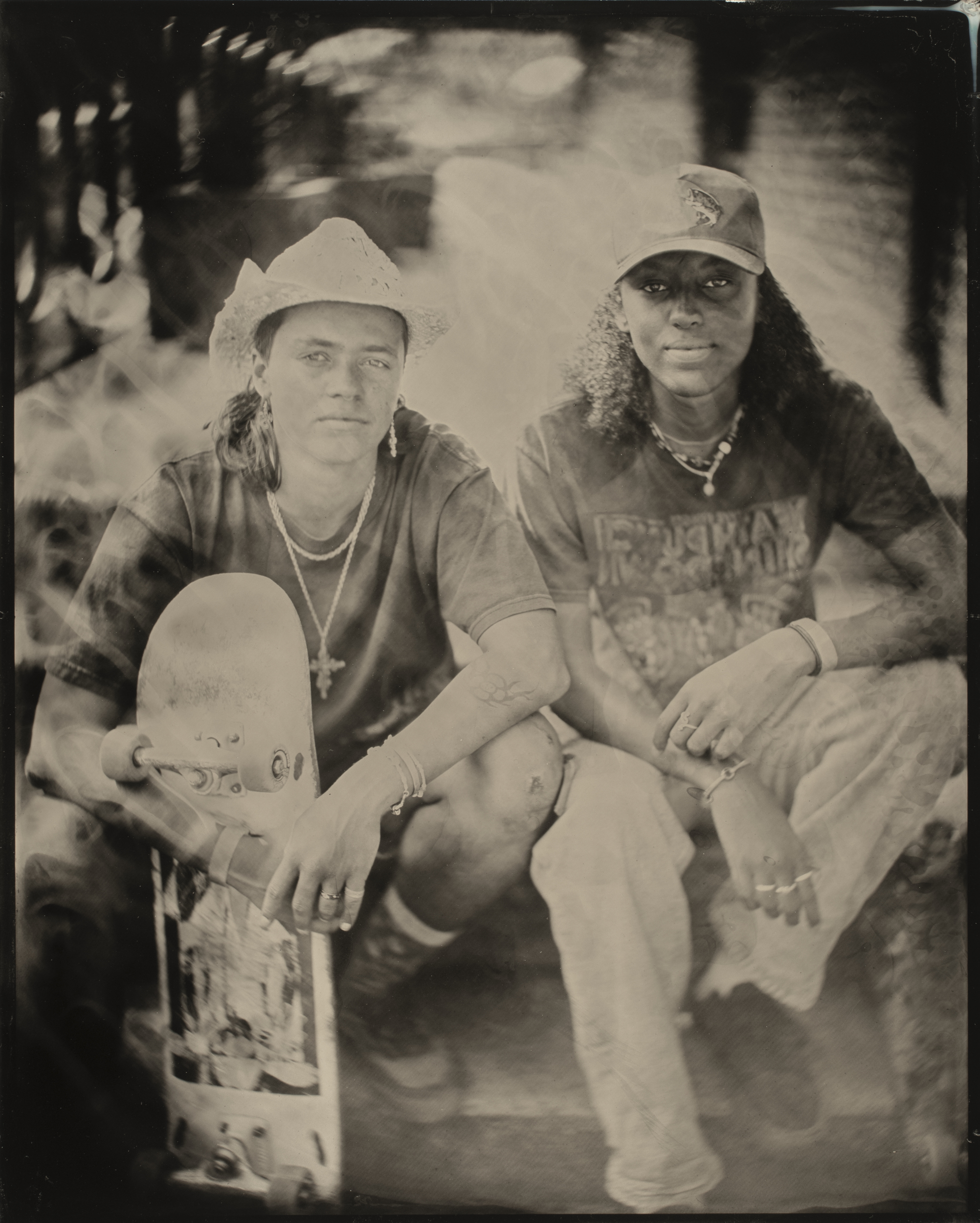Two individuals are sitting close together; one wears a cowboy hat and holds a skateboard, while the other sports a cap. The image has a vintage, sepia tone.