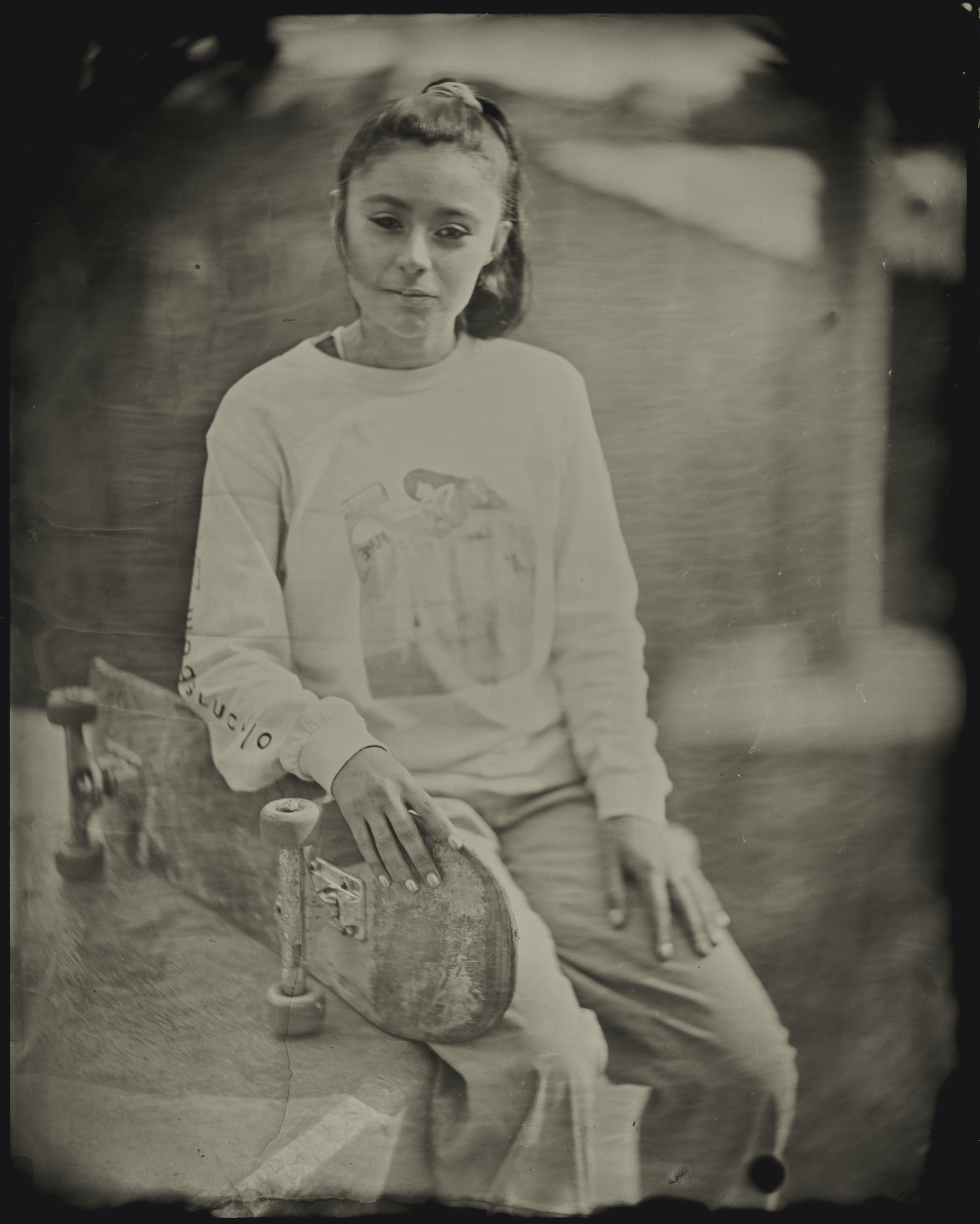 A person with long hair sits holding a skateboard. They're wearing a sweater with a graphic design. The image has a vintage, sepia tone.