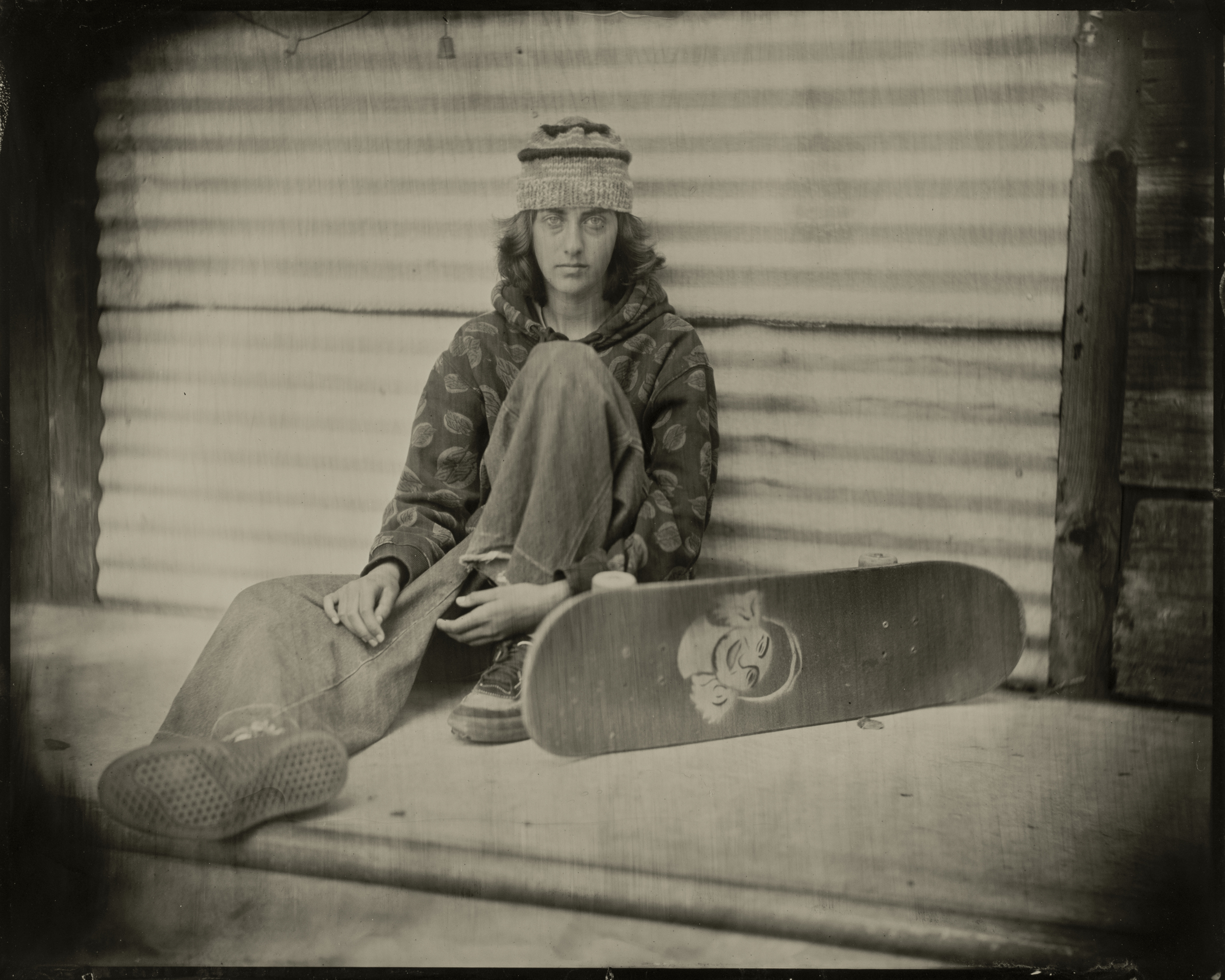 A person sits casually, wearing a beanie and patterned hoodie. They lean against a corrugated wall, with a skateboard featuring a face graphic beside them.