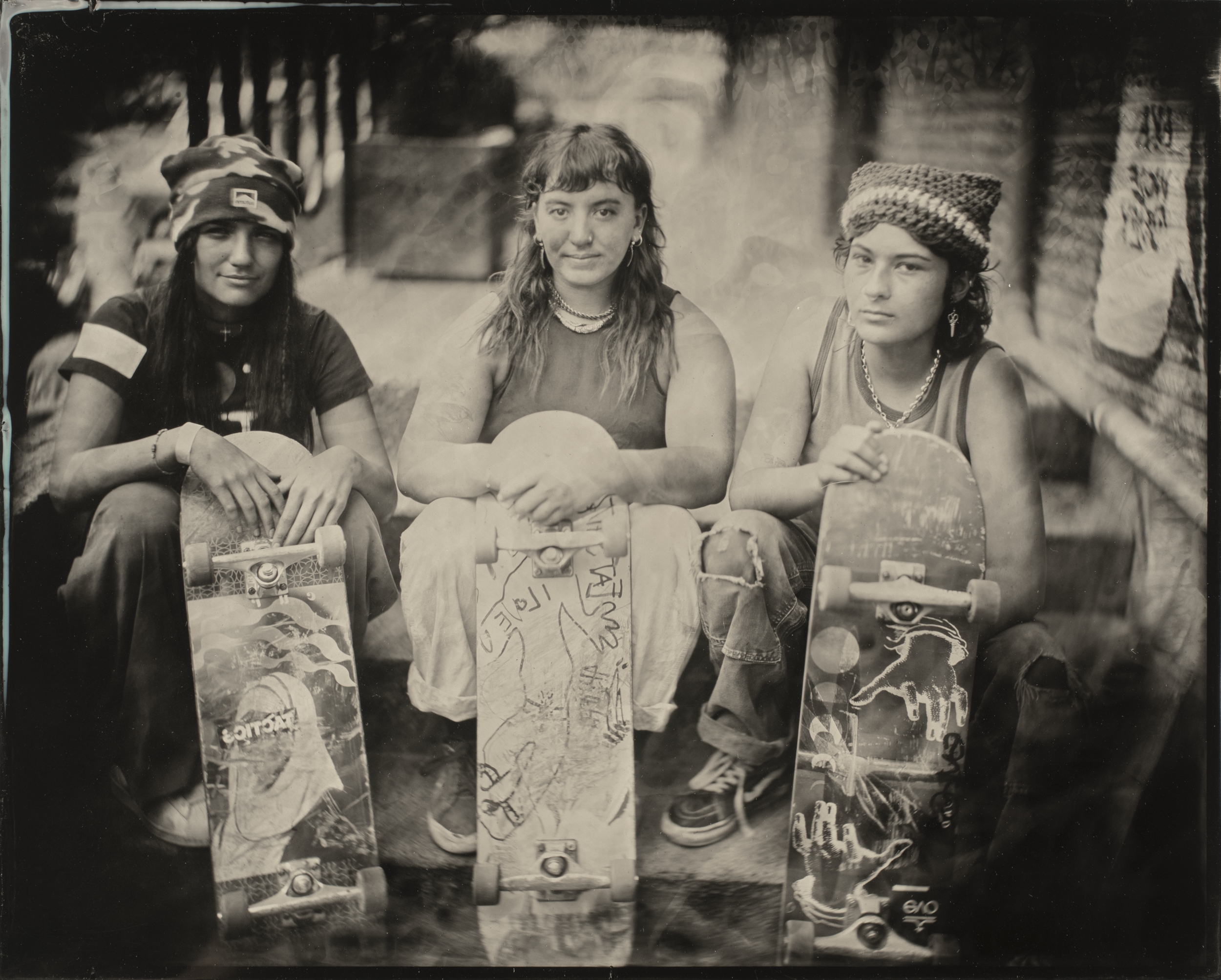 Three people sit side by side, each holding a skateboard vertically. They are dressed casually and look directly into the camera. The image is in black and white.