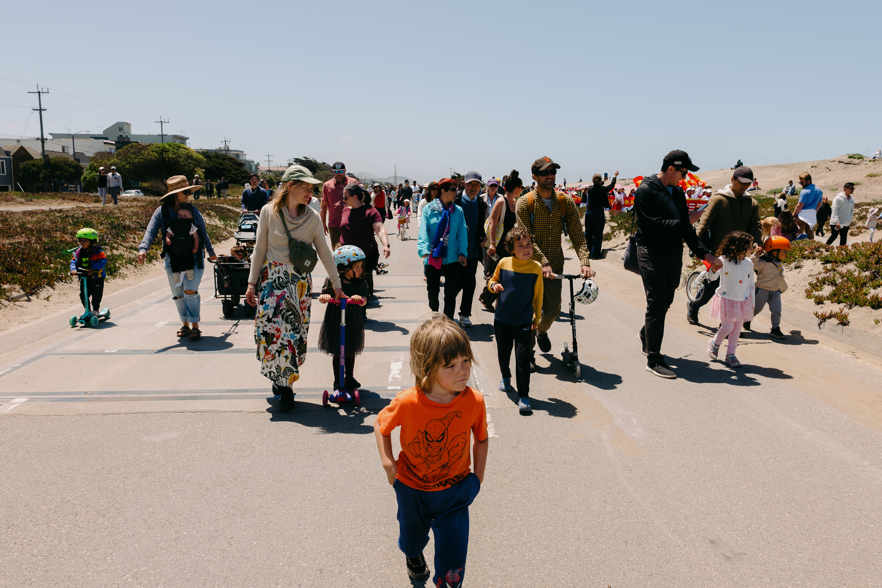 A lively group of people, including children on scooters and adults, walk along a sunny path by a sandy landscape. The scene is busy and vibrant with activity.