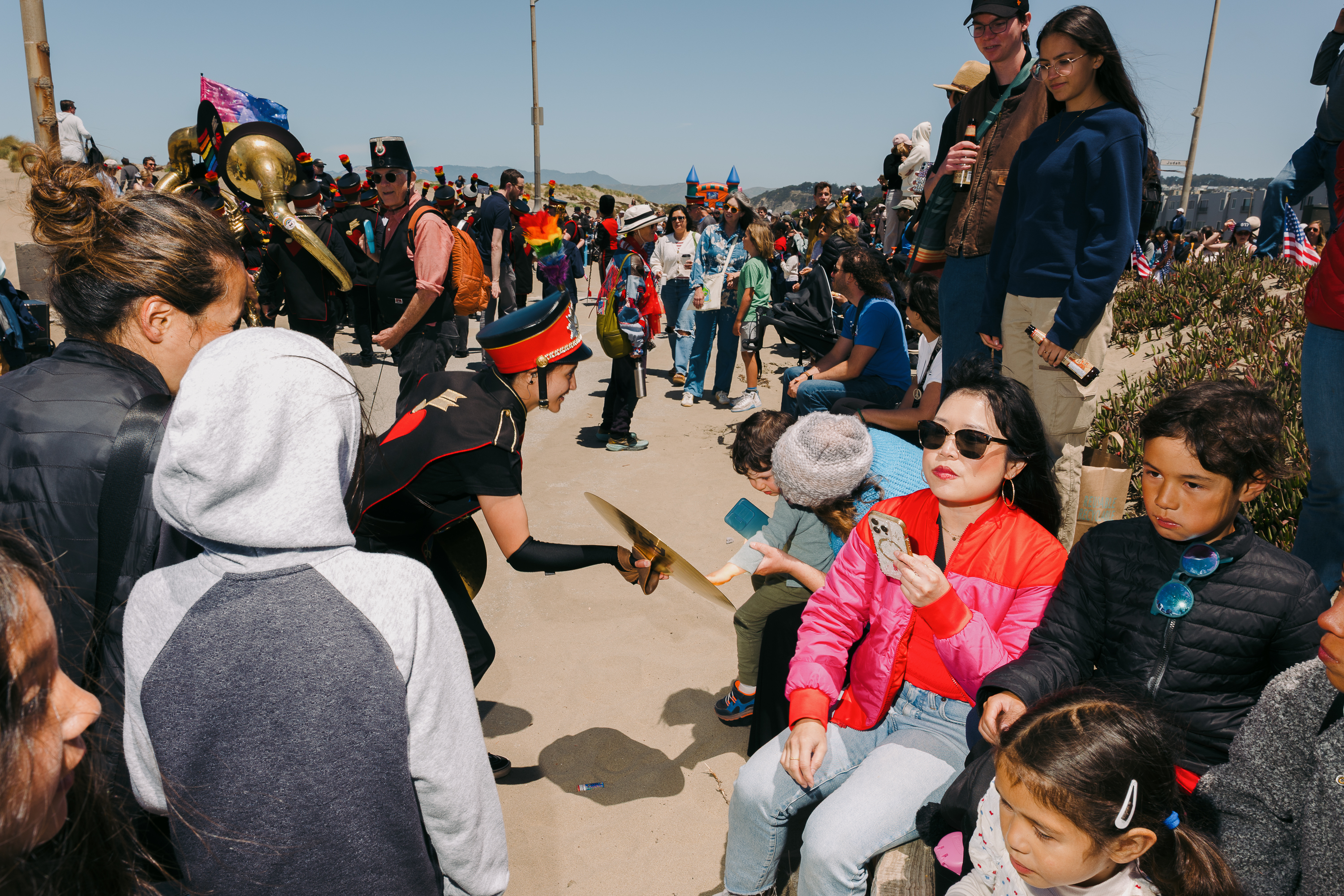 A marching band performs outdoors on a sunny day, surrounded by people. A band member in uniform is playing cymbals. Spectators, some seated, are observing.