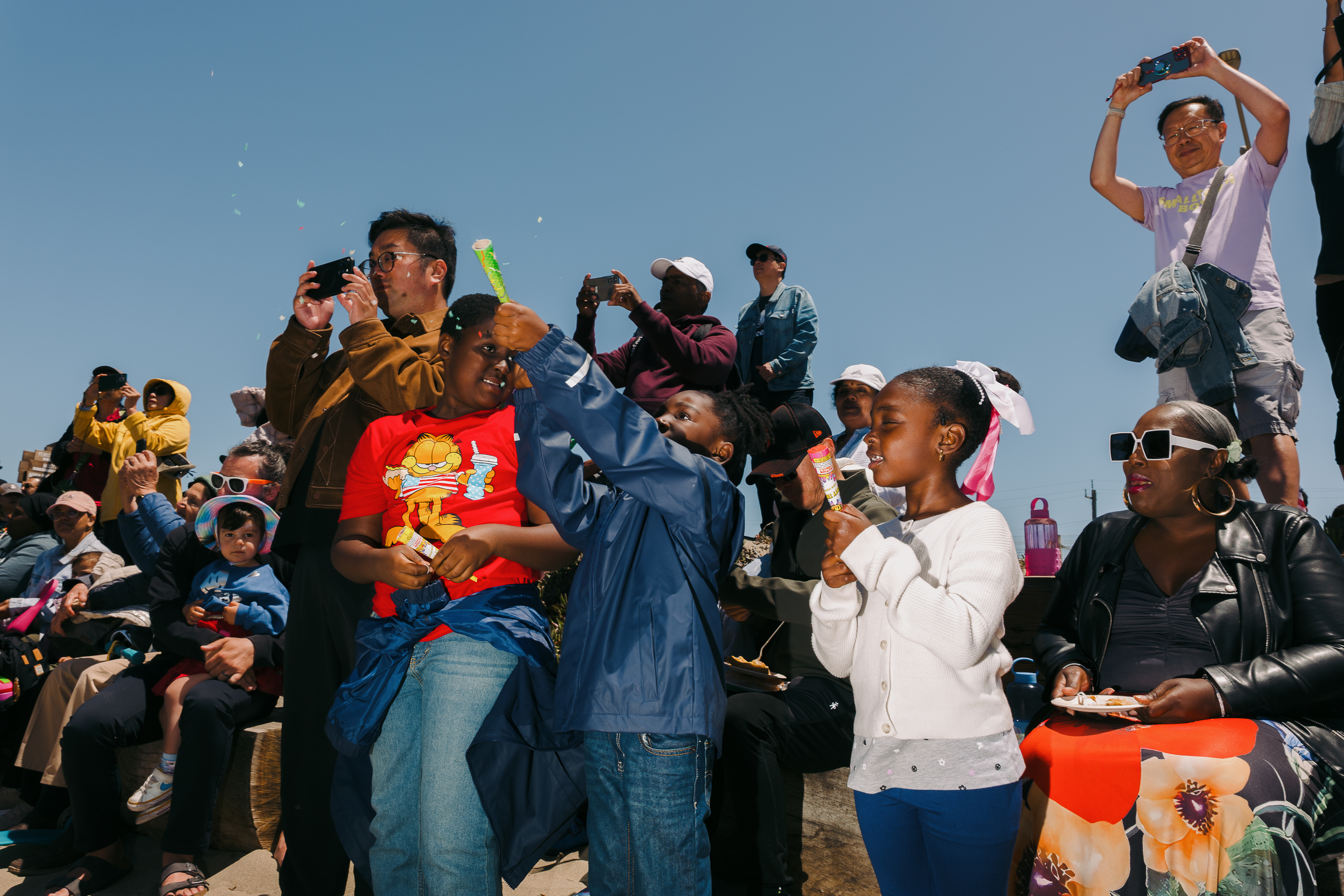 A diverse group of people, including children and adults, are gathered at an outdoor event. Some are blowing bubbles, others taking photos, all appear to be enjoying themselves under a clear blue sky.