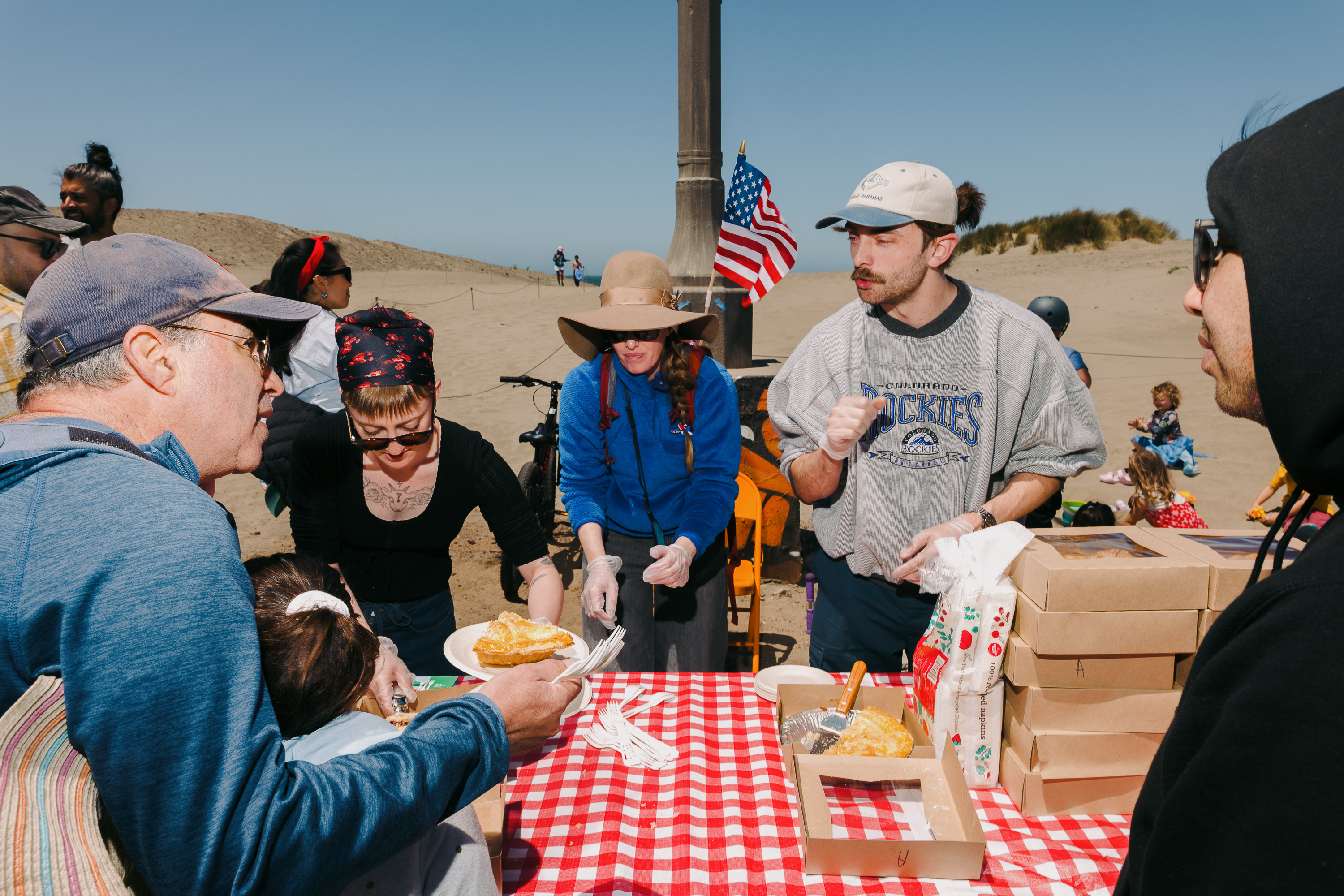 A group of people stands at a red-checkered table on a beach, distributing food with an American flag in the background.