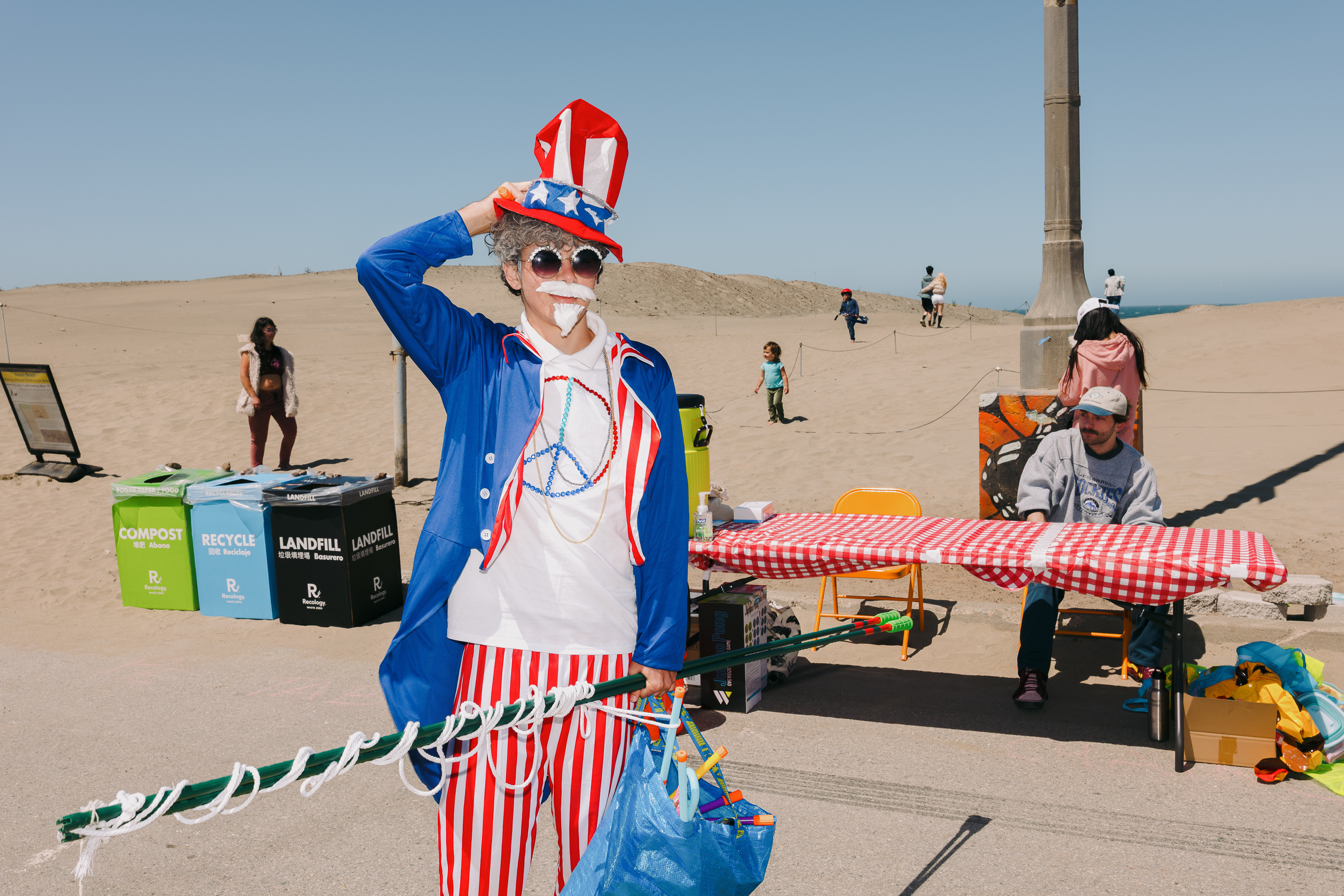 A person dressed in a festive red, white, and blue costume stands on a sandy area with recycling bins and a table with a checkered cloth nearby.