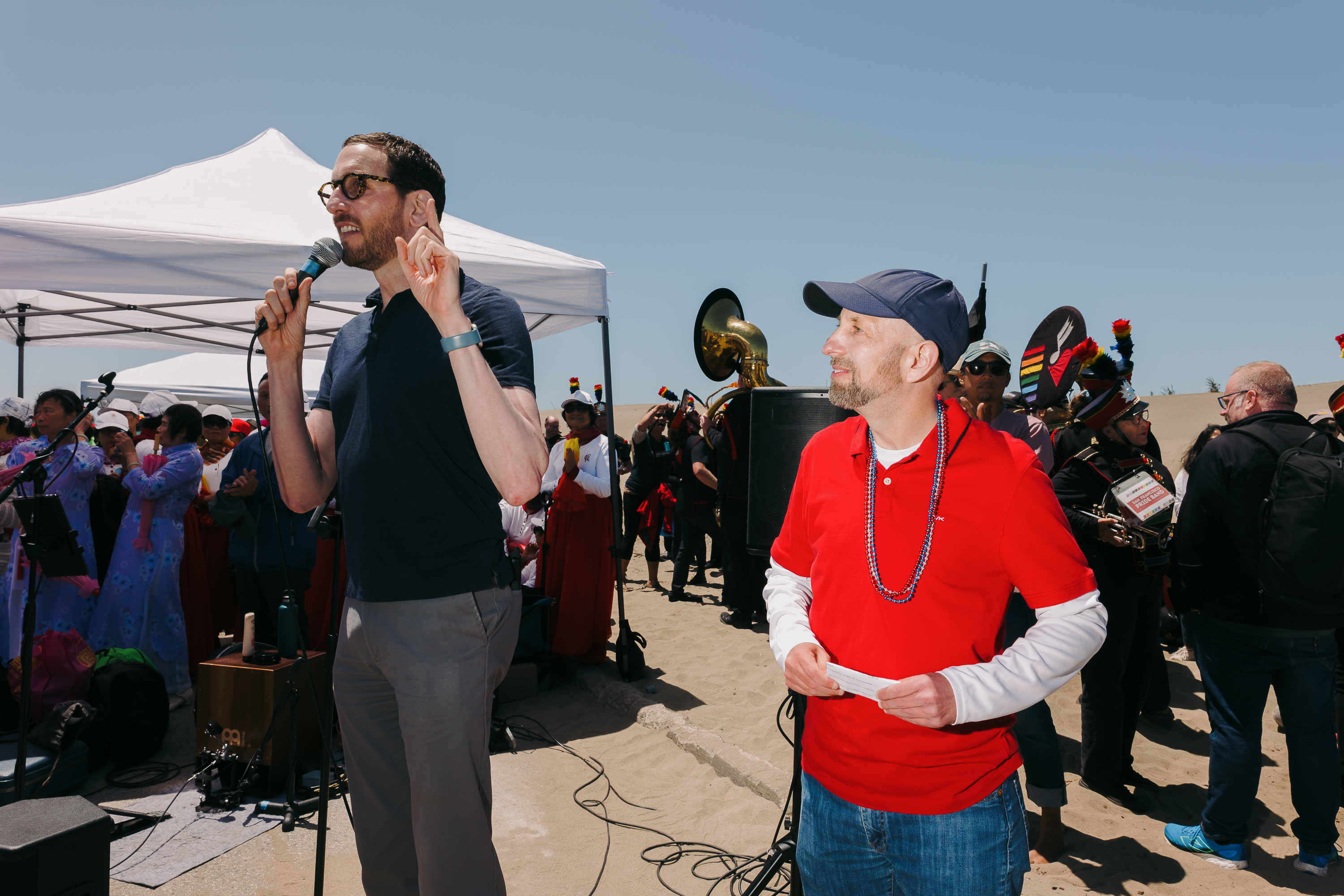 Two men stand outdoors, one speaking into a microphone. They're surrounded by a crowd, with a band playing instruments nearby. A white tent is visible.