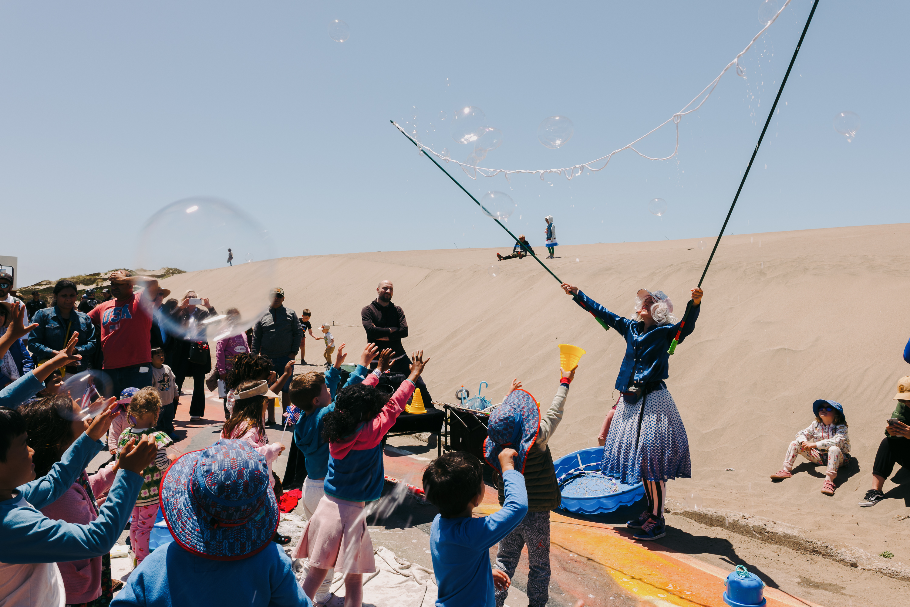 A group of children eagerly reach for large, floating bubbles created by a person using poles, against a sandy background under a clear blue sky.