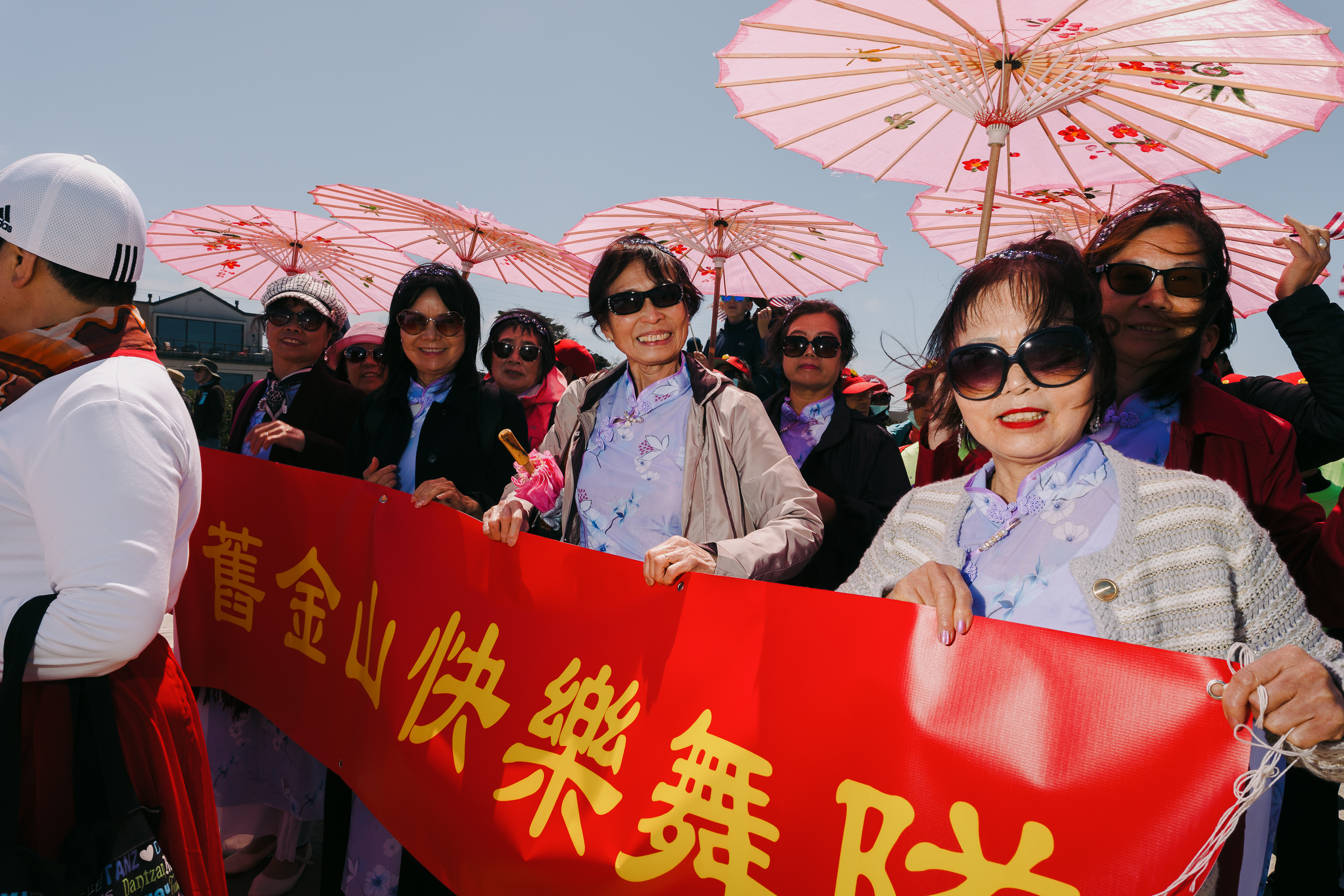 A group of smiling women wearing sunglasses and floral attire hold pink parasols and display a red banner with yellow Chinese characters, standing under a clear sky.