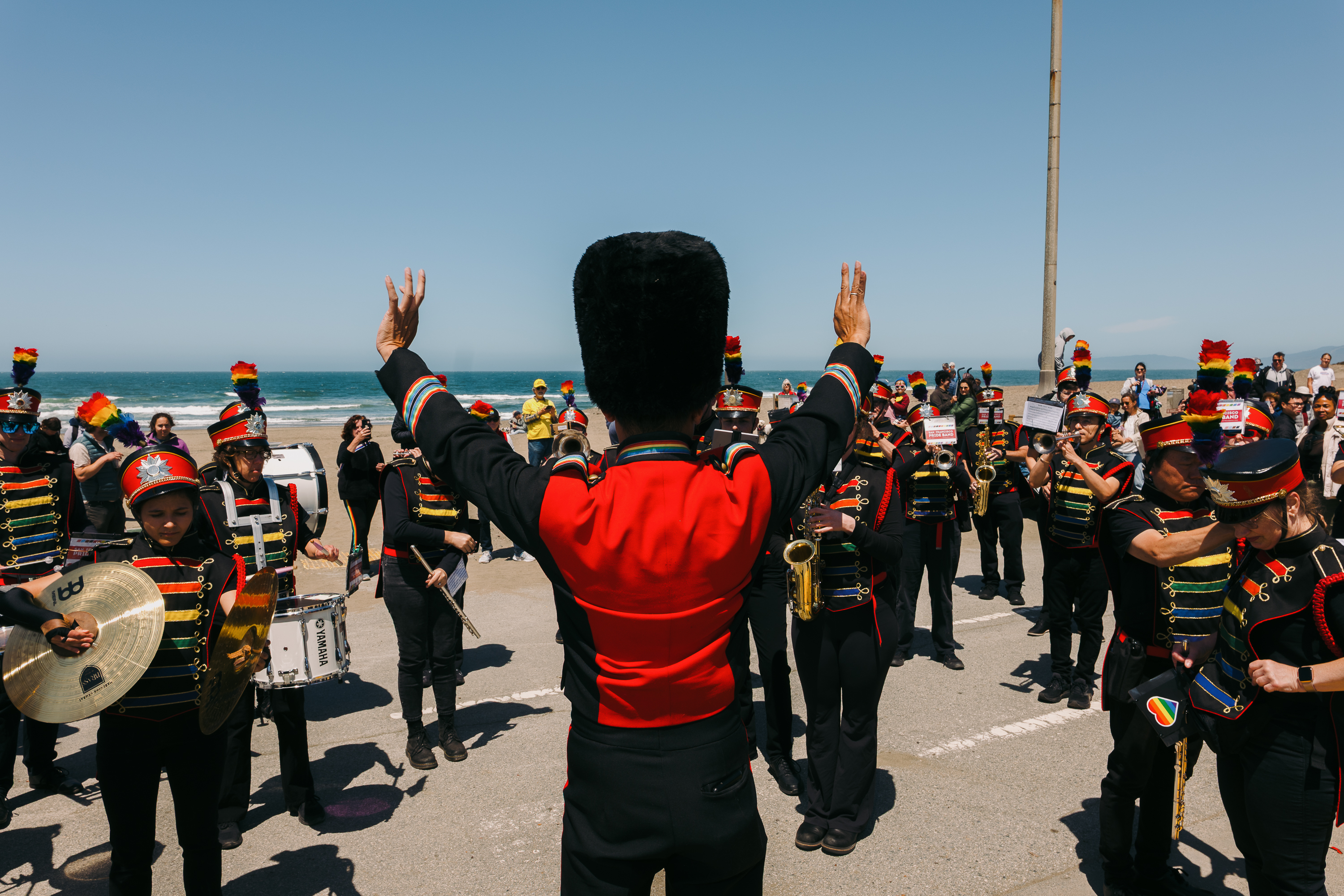 A marching band in colorful uniforms plays on a seaside street as spectators watch. The conductor stands with raised arms facing the musicians.
