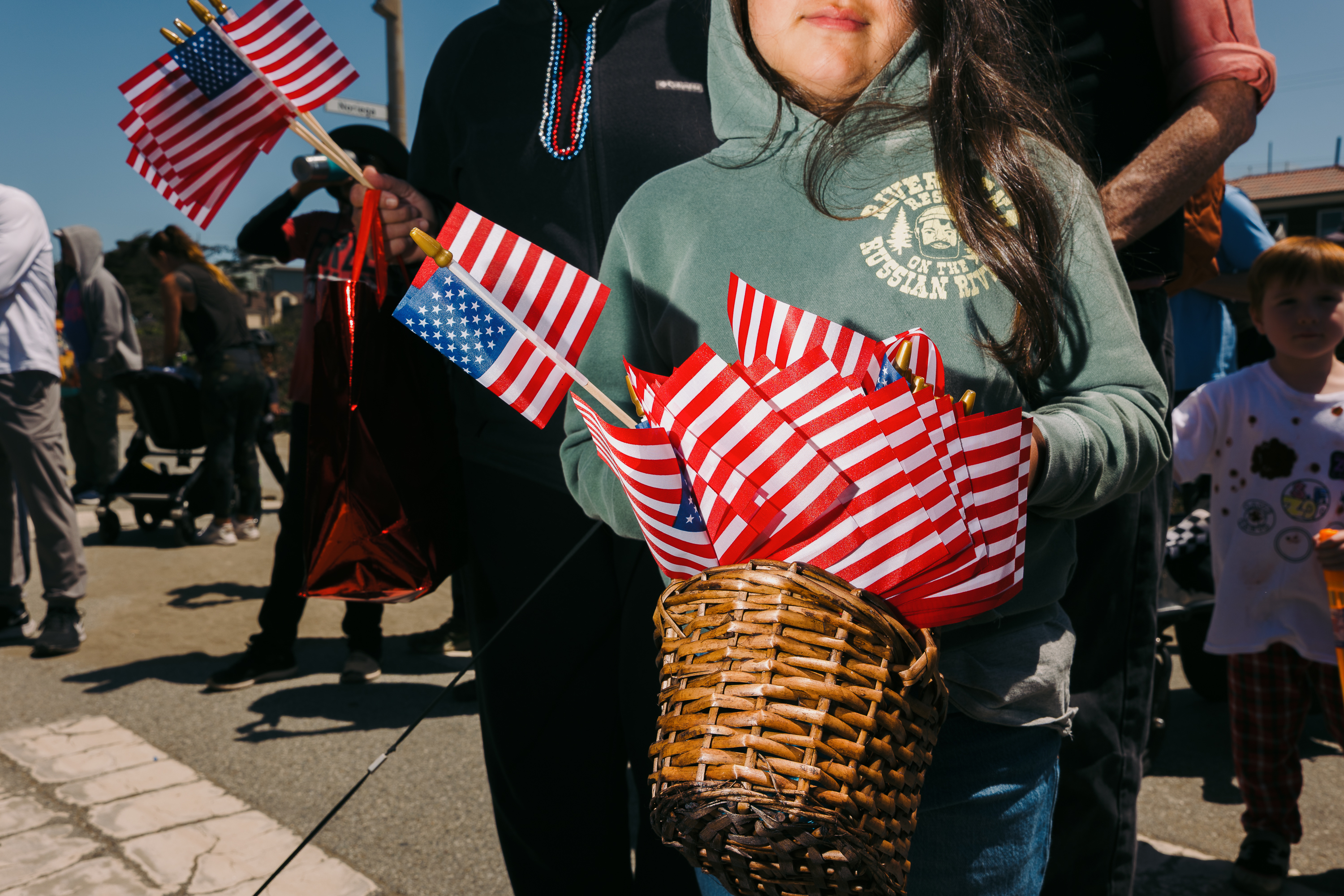 A person in a green hoodie holds a basket full of small American flags, standing among a crowd. A child and other people with strollers are in the background.