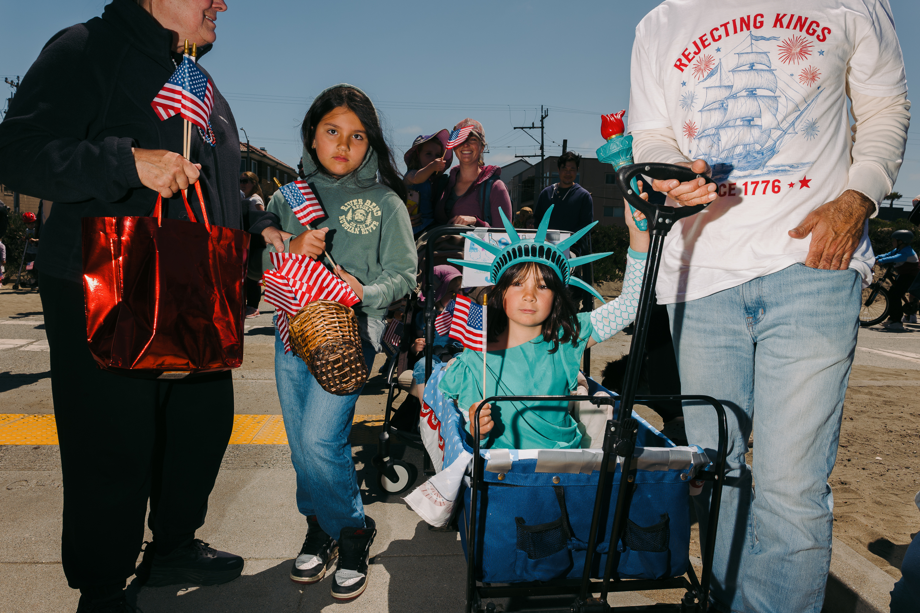 A child in a Statue of Liberty costume holds a small American flag in a wagon, surrounded by people with flags and festive attire on a sunny day.