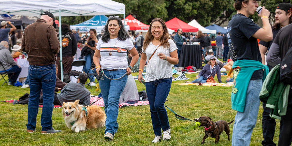 Two women walk dogs at a lively outdoor event. People sit on blankets, tents are in the background, and there's a cheerful, bustling atmosphere.