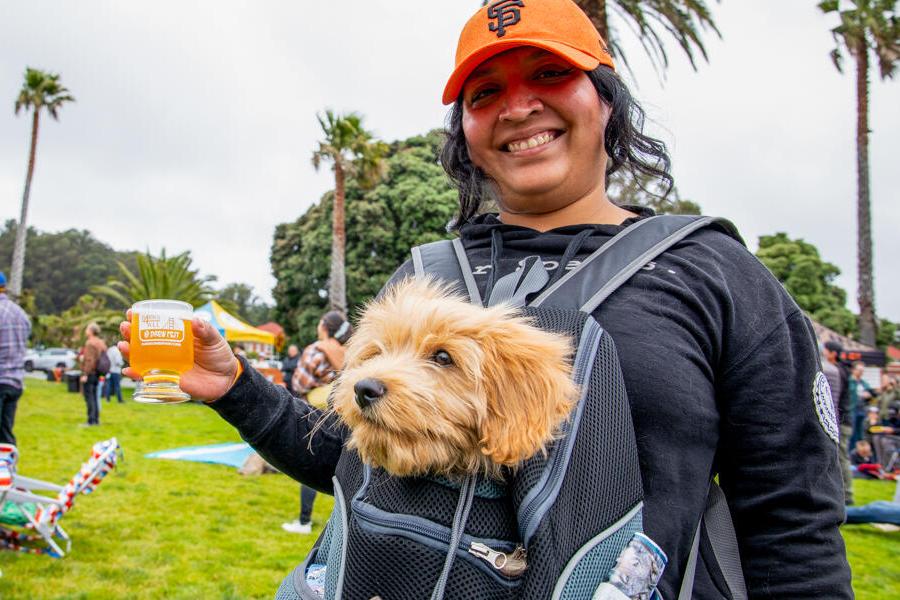 A smiling person in an orange cap holds a drink. A small dog is in a front carrier. They're outdoors with people and palm trees in the background.