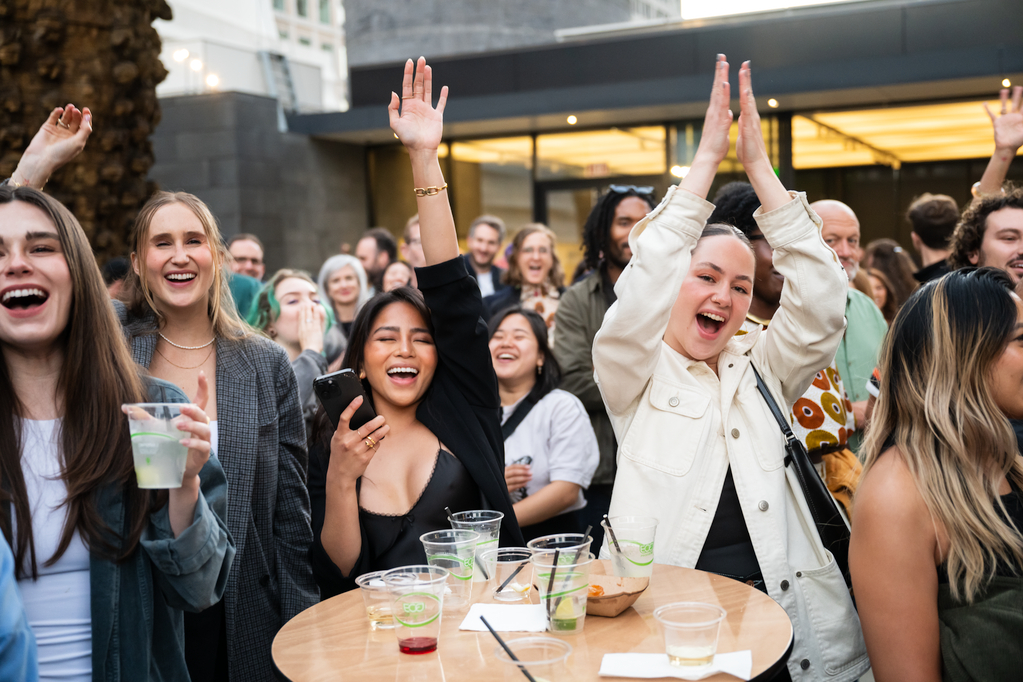 A lively group of people at an outdoor event are smiling, cheering, and holding drinks, with a woman in a white jacket raising her hands in excitement.