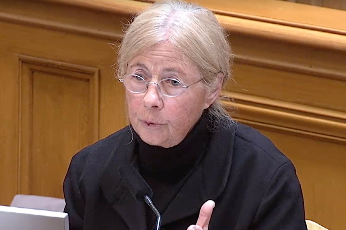 A person with light hair and glasses is speaking, seated indoors. They are wearing a black jacket and are positioned in front of a laptop next to a wooden wall.