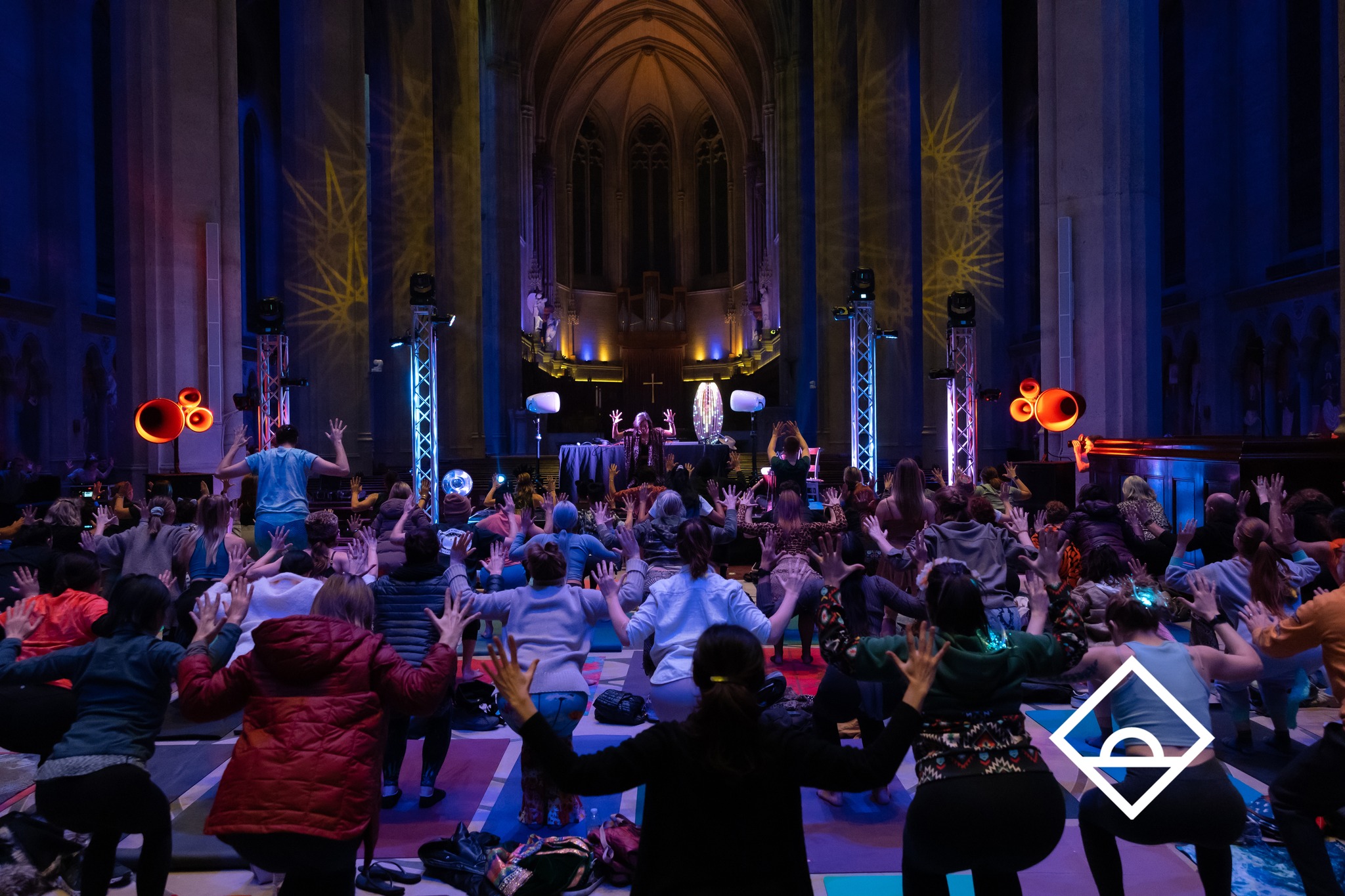A large group is participating in a yoga or dance session inside a dimly lit, grand hall, with colorful lights and elevated speakers enhancing the atmosphere.