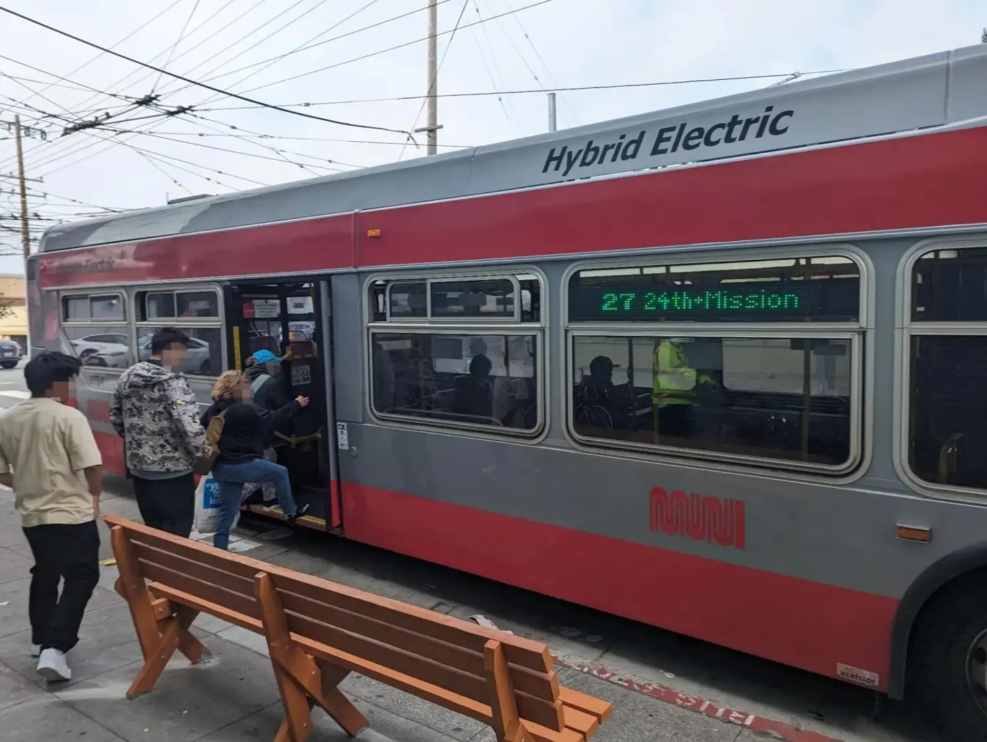 People are boarding a gray and red hybrid electric bus labeled &quot;27 24th Mission&quot; at a bus stop. There are benches and overhead wires visible.
