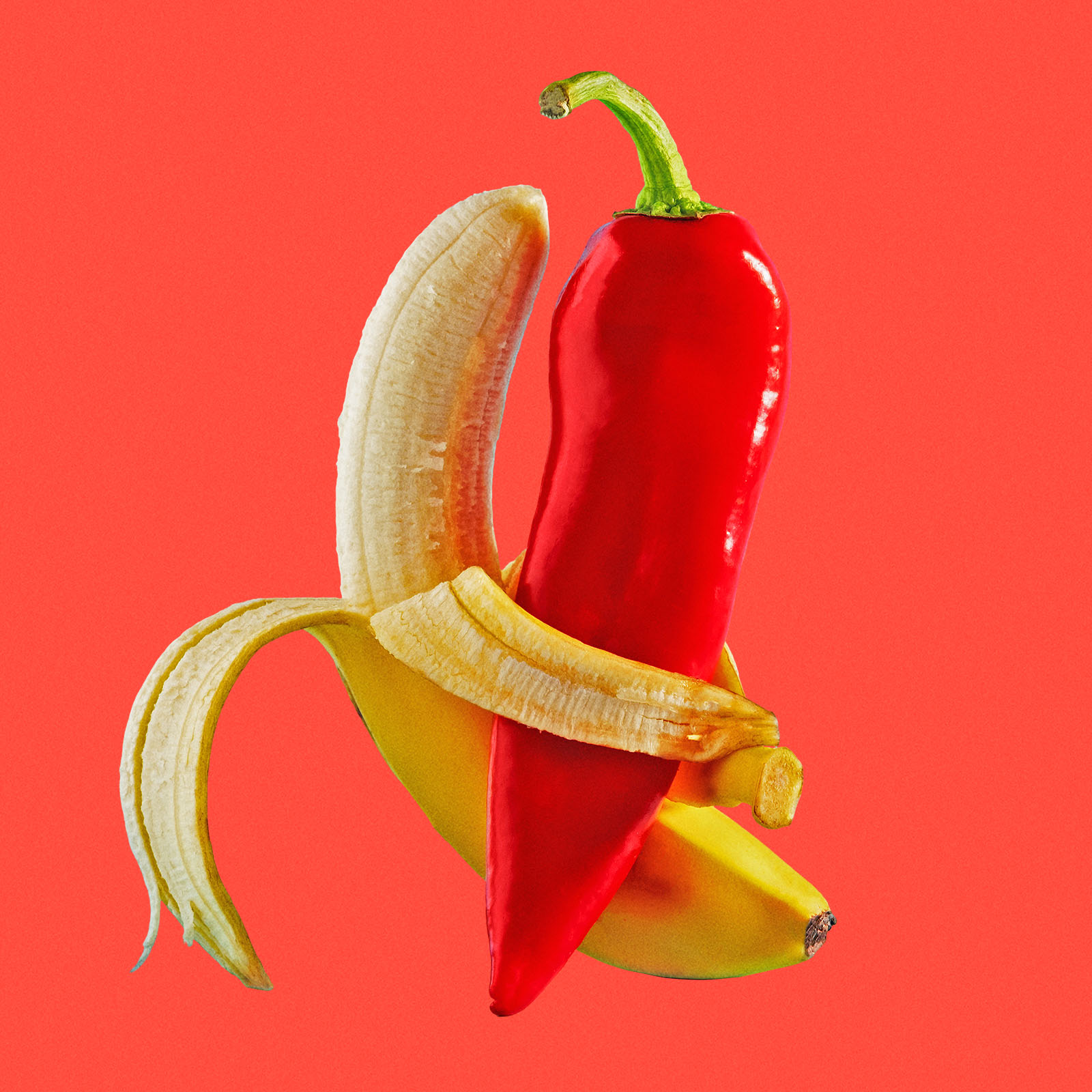 A peeled banana is wrapped around a bright red chili pepper on a red background, creating a striking, surreal visual contrast.