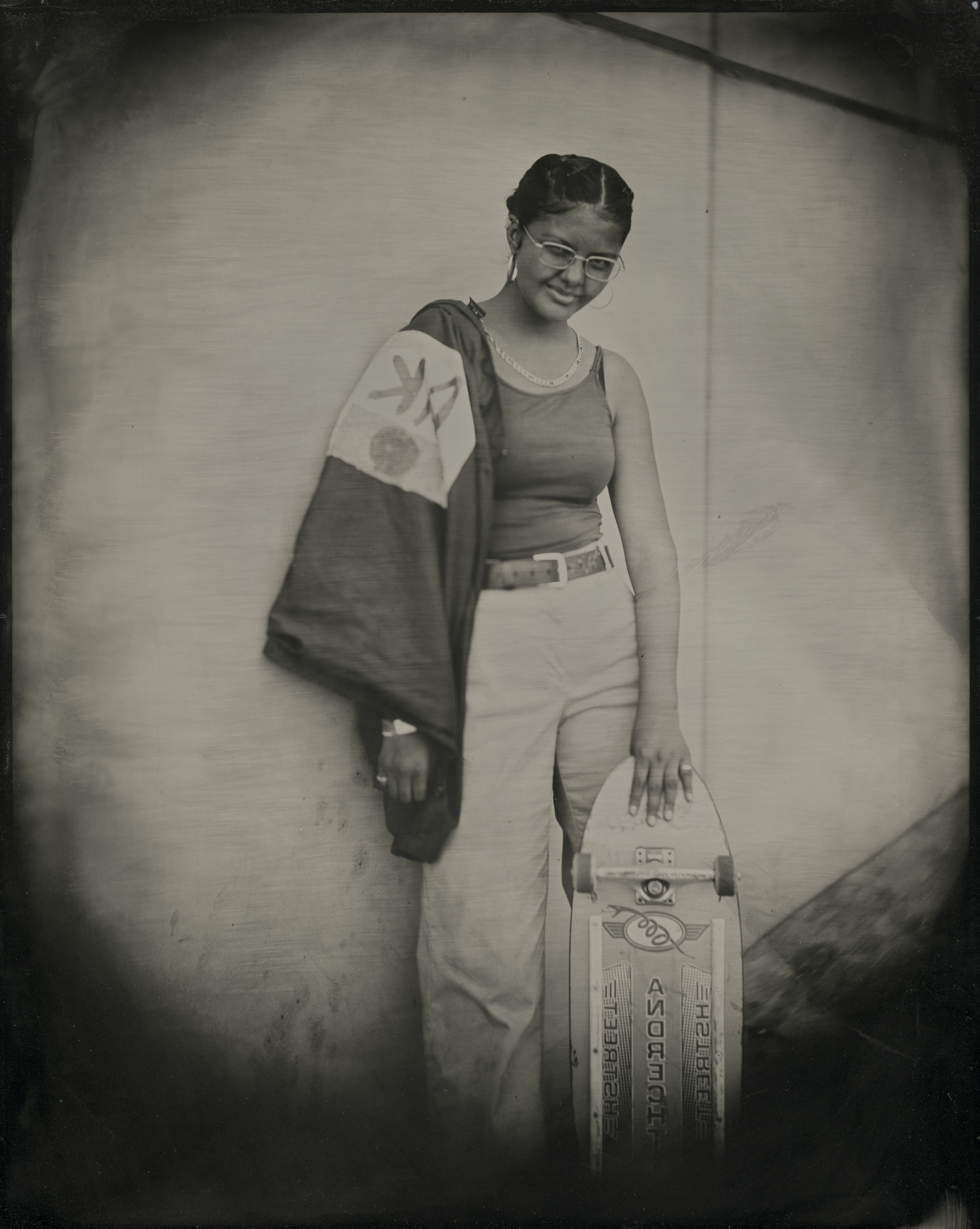 A person leans against a wall, smiling. They wear glasses, a tank top, light pants, and hold a skateboard with one hand while a jacket drapes over their shoulder.