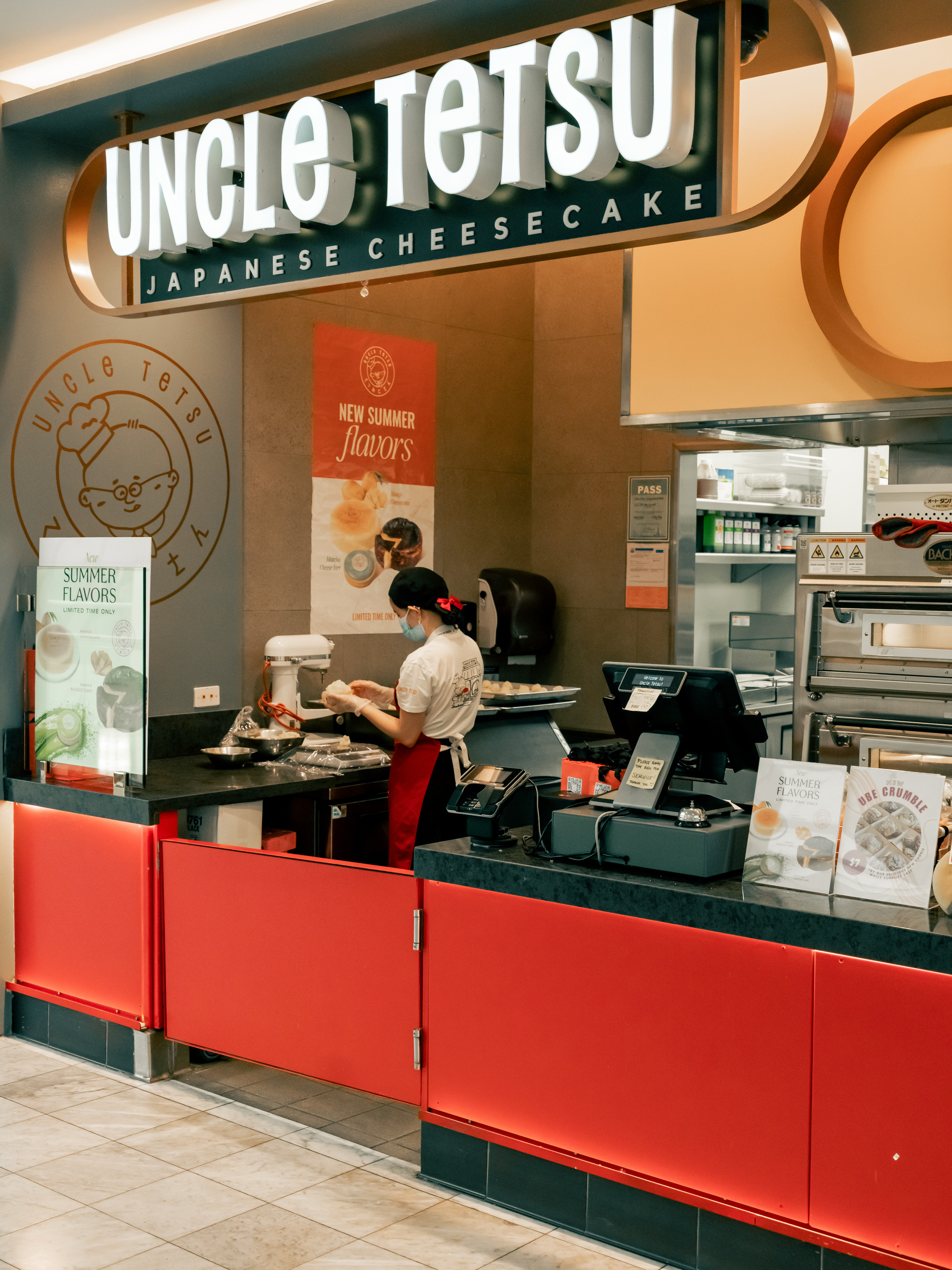 A bakery counter with a sign reading &quot;Uncle Tetsu Japanese Cheesecake.&quot; A person is working behind the counter with signage for summer flavors displayed.