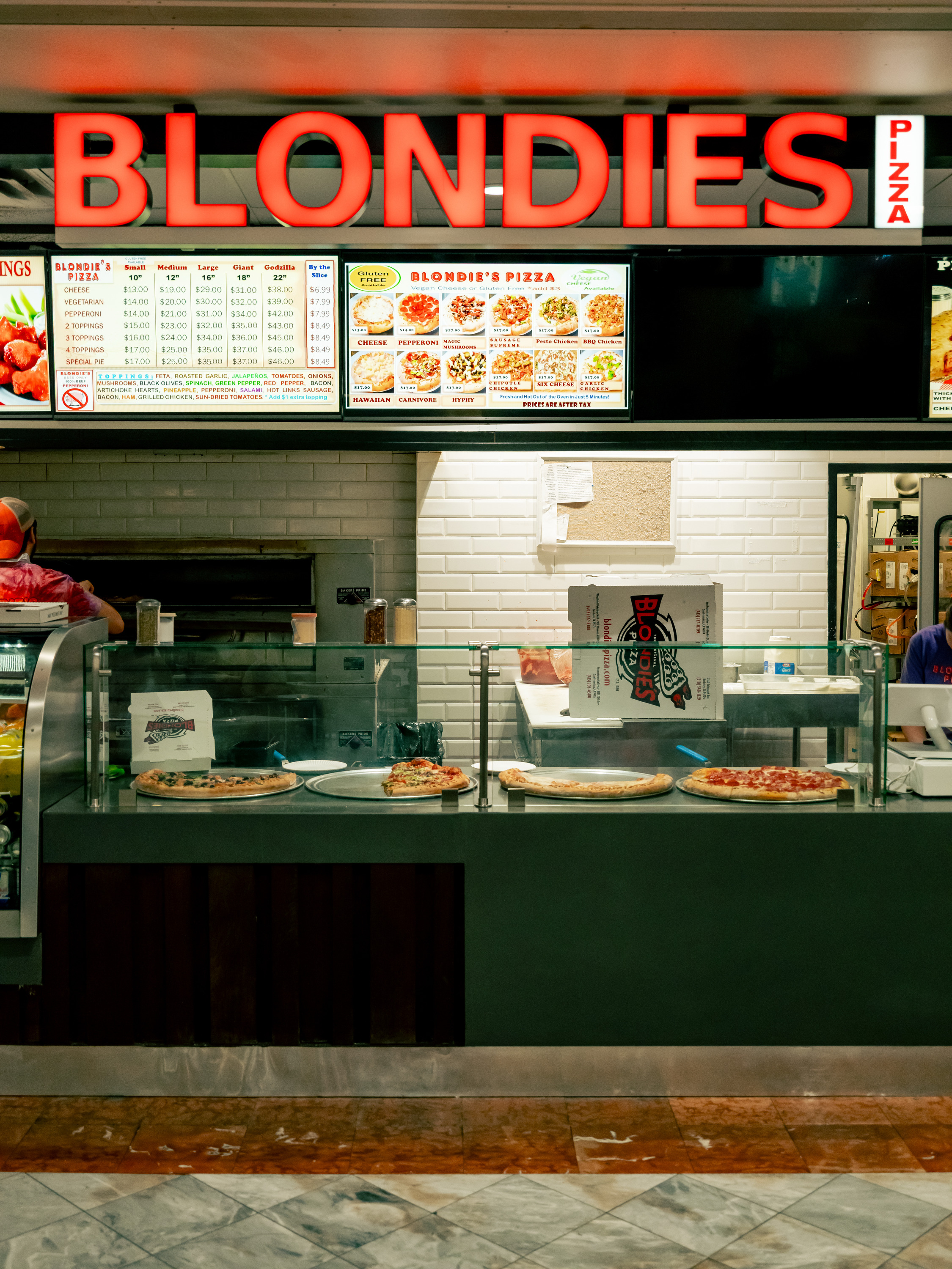 A pizza shop called &quot;Blondies&quot; shows an illuminated sign, menu boards, and pizzas on display behind a glass counter in a tiled, indoor setting.