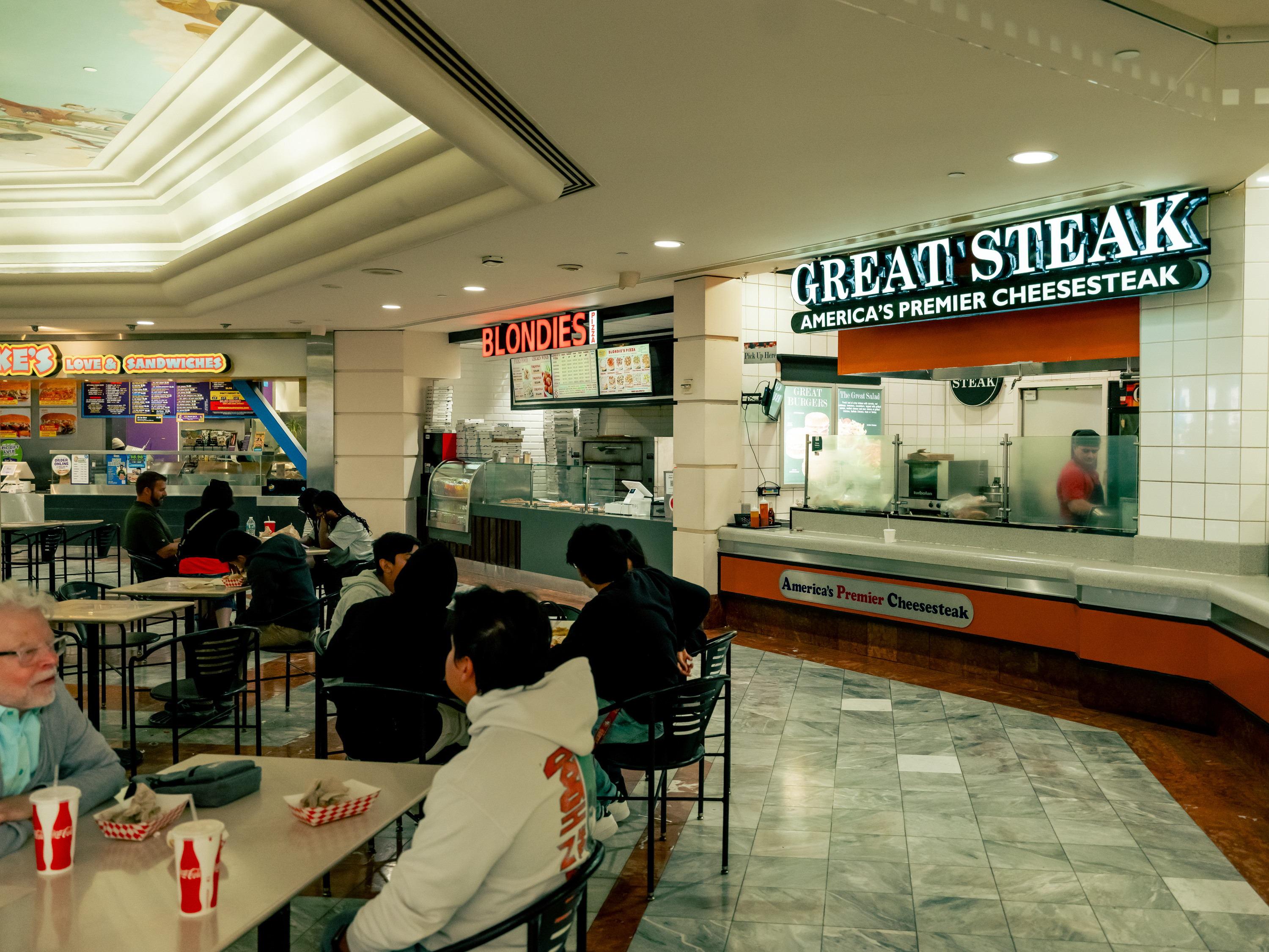 A food court scene with people seated at tables. Fast food outlets, including &quot;Blondies&quot; and &quot;Great Steak,&quot; are visible in the background.