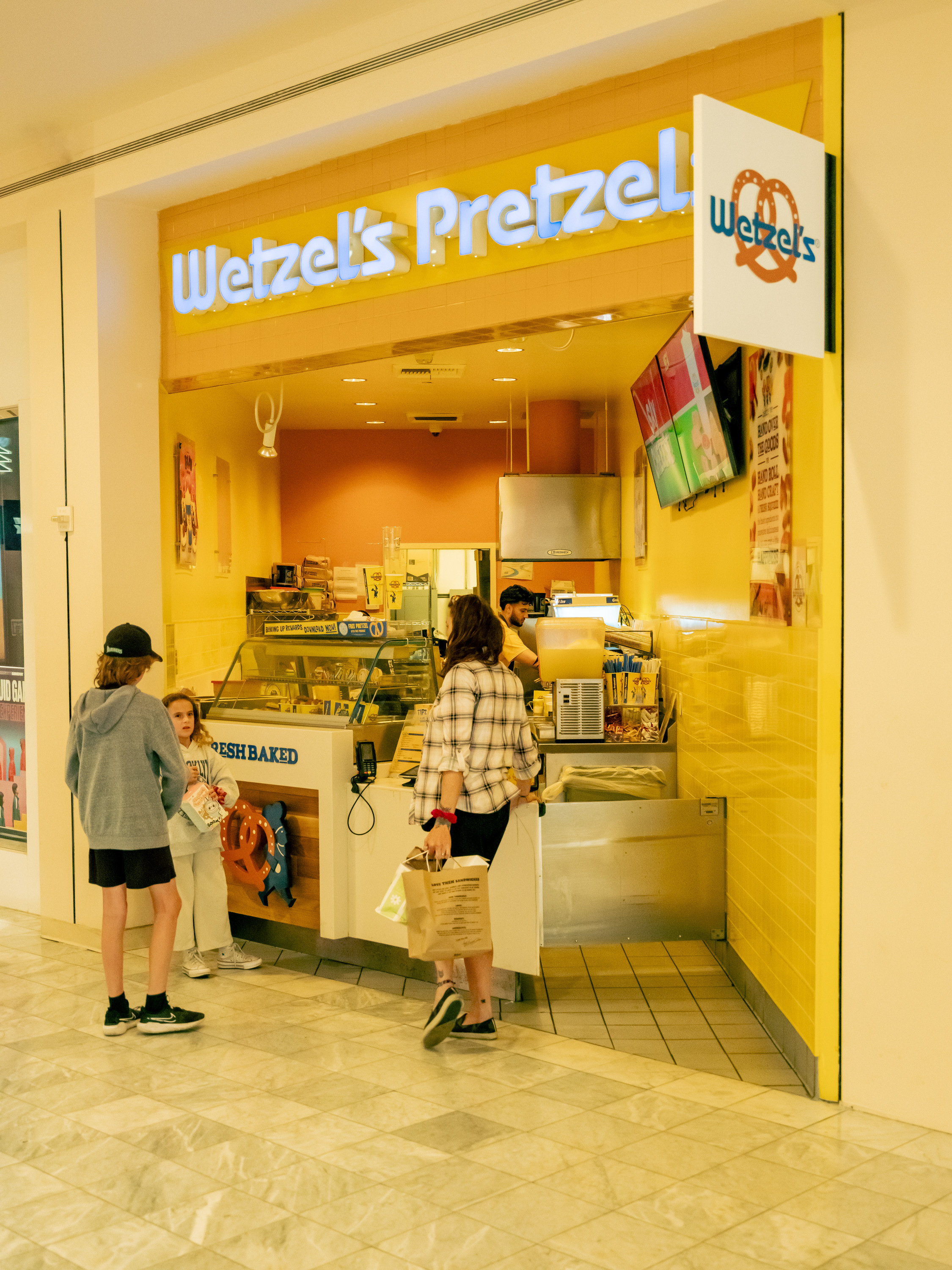A Wetzel’s Pretzels store in a mall features a bright yellow interior. People are in line to order, and a worker is behind the counter preparing food.