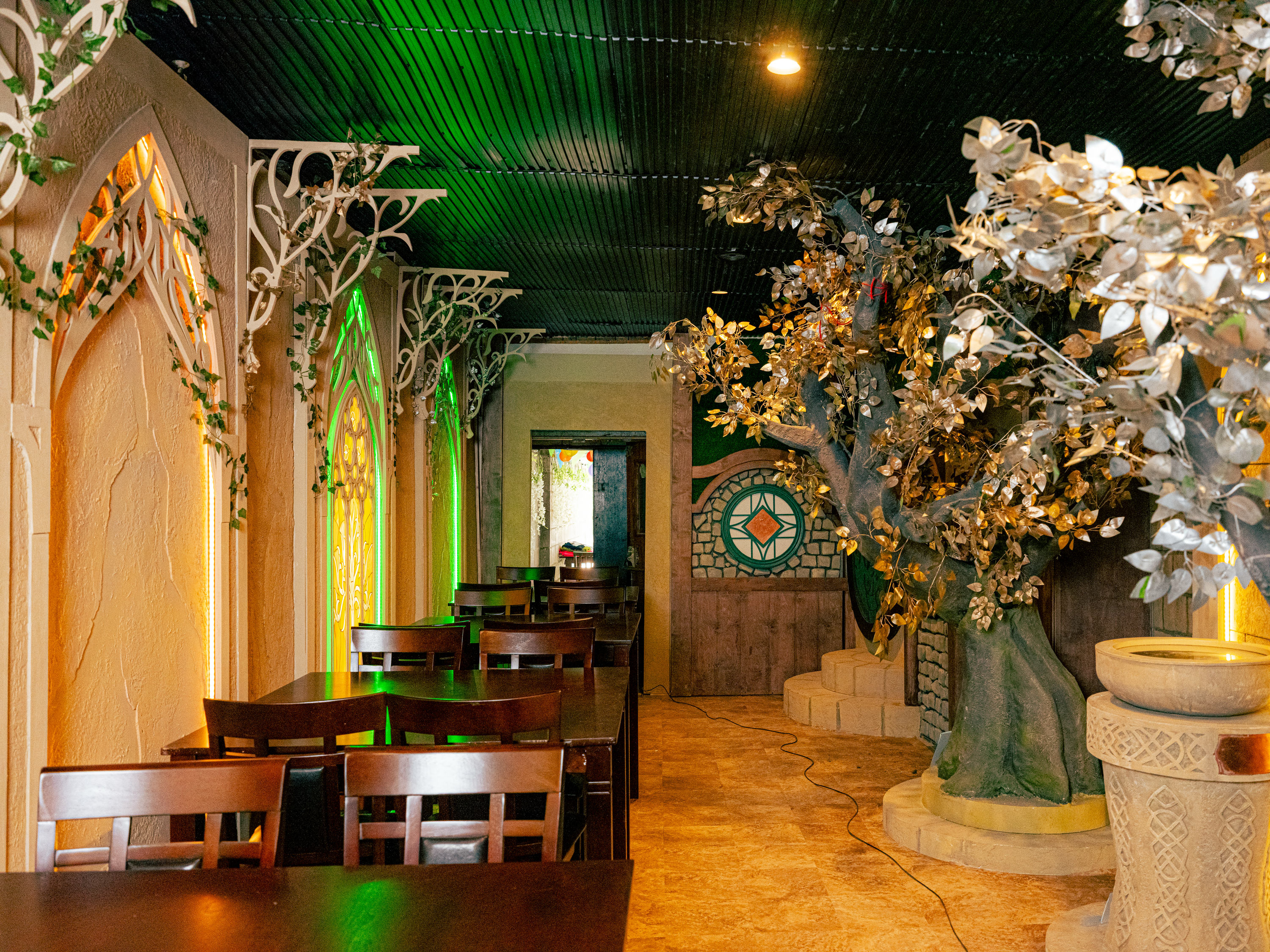 The image shows a fantasy-themed room with wooden tables and chairs, a faux tree with metallic leaves, arched windows with greenery, and intricate wall designs.