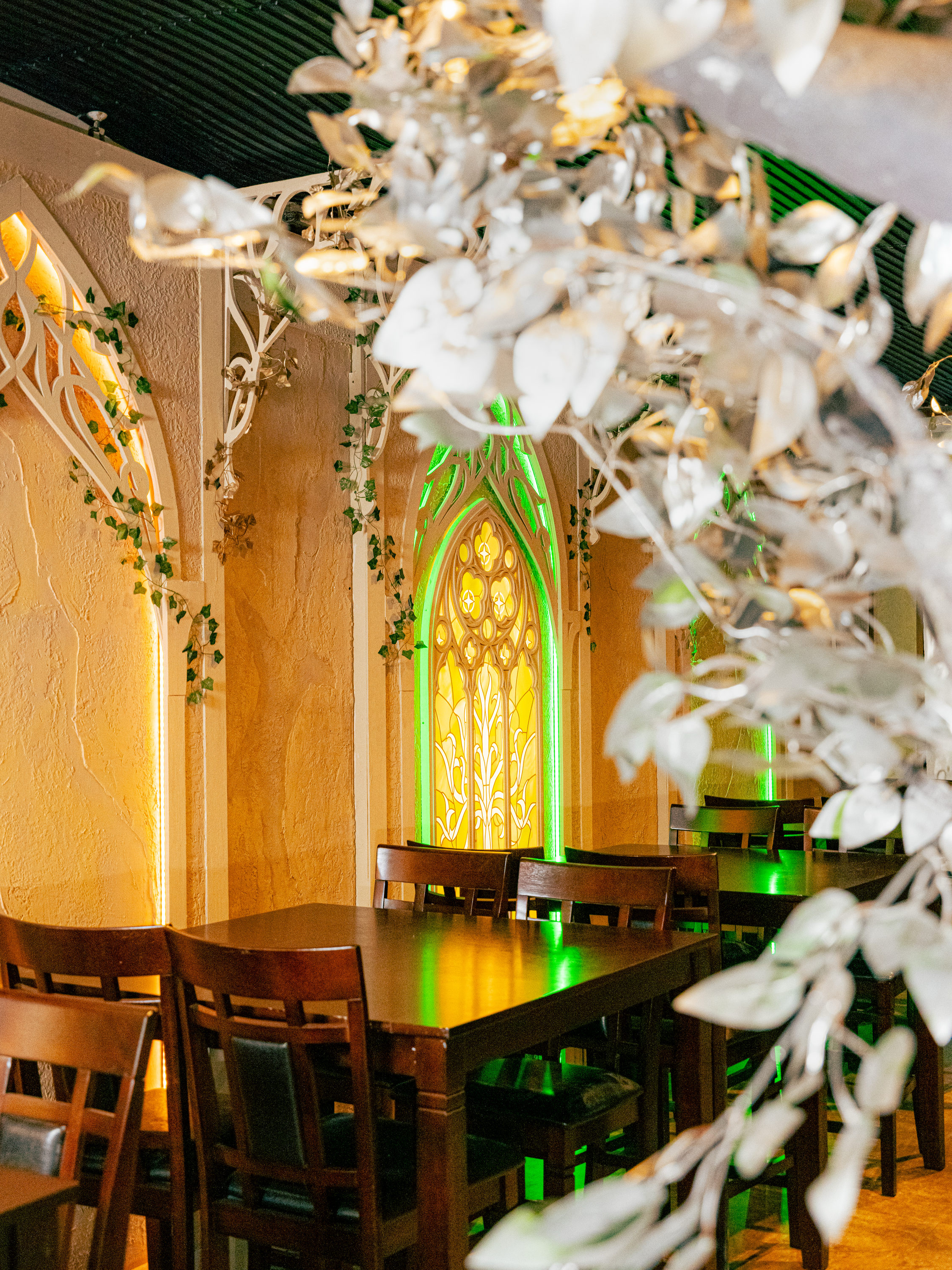 The image shows a cozy room with wooden tables and chairs, decorated with green and yellow stained glass windows, leafy vines, and warm lighting.