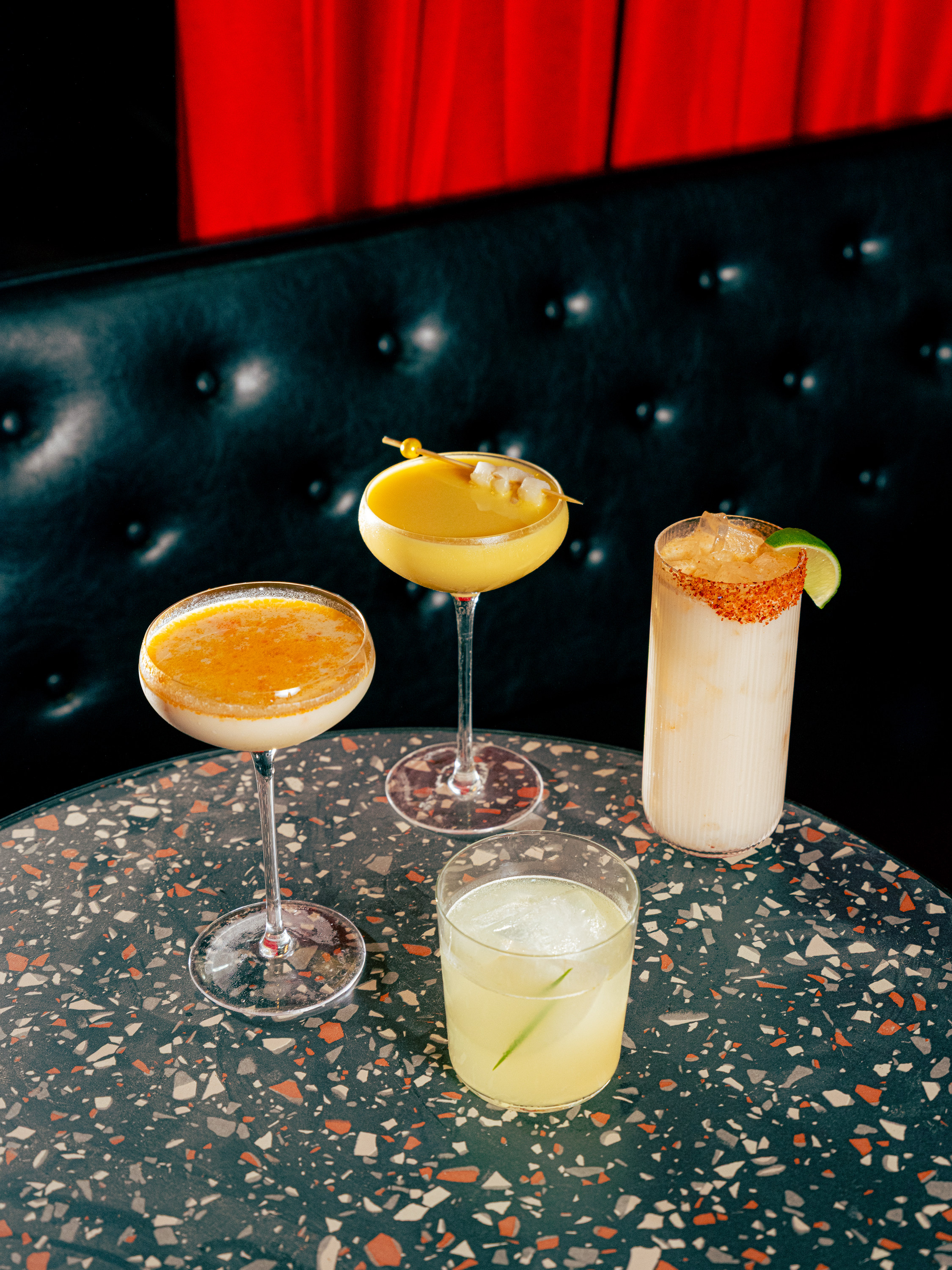 Four colorful cocktails are on a terrazzo table: two in coupe glasses, one in a tall glass with a lime wedge, and one in a short glass with ice. A red curtain and tufted black booth are in the background.