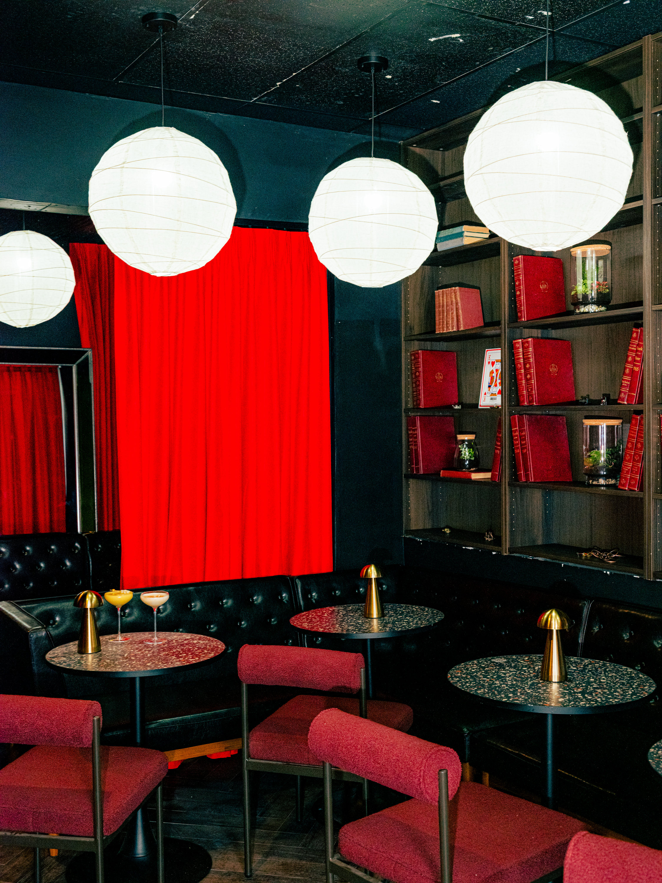 The image shows a cozy room with red chairs and tables, pendant lights, a black leather couch, red curtains, and bookshelves filled with red books.