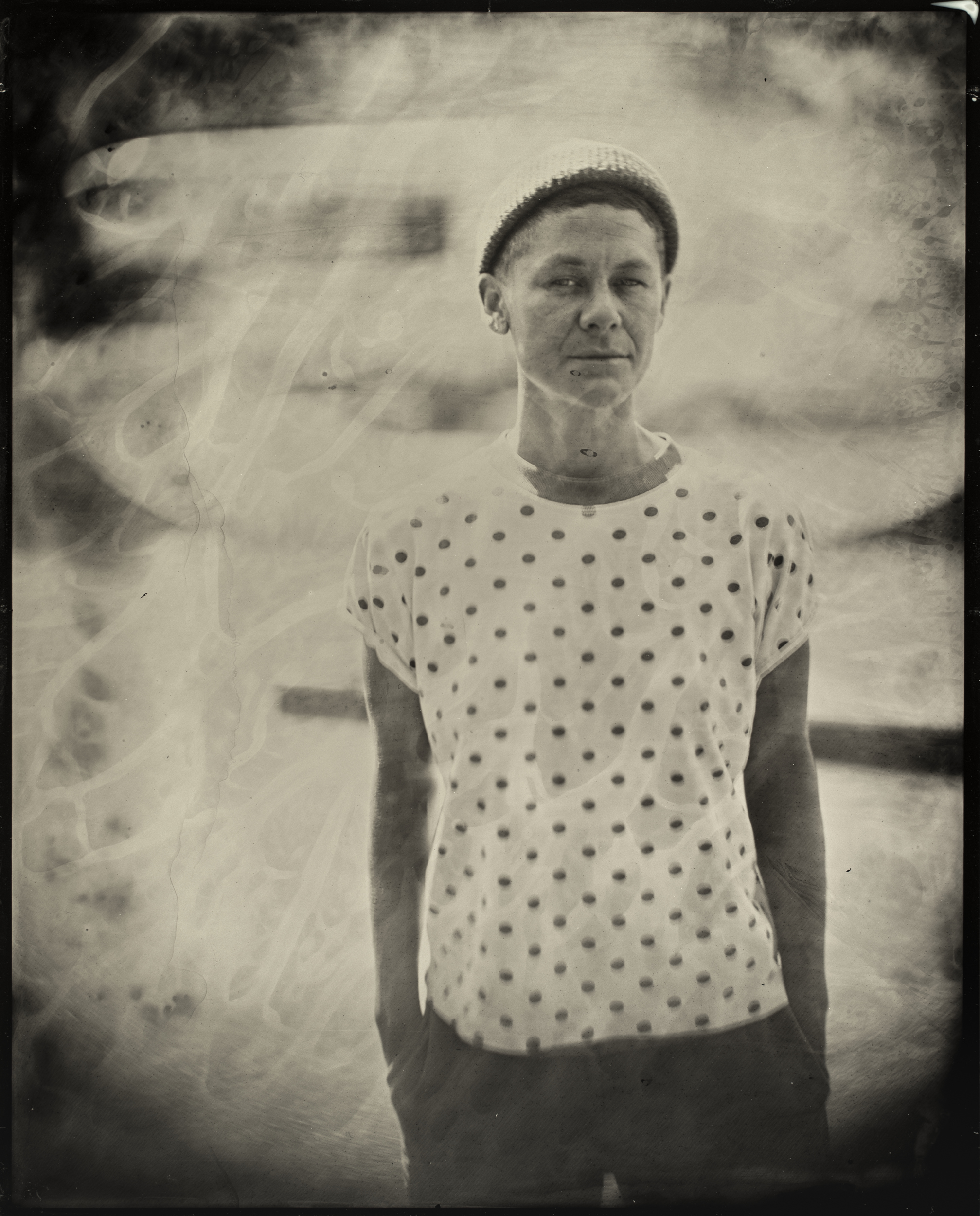 A person stands with a neutral expression, wearing a polka dot shirt and a beanie. The image has a vintage, sepia-toned effect with soft focus.
