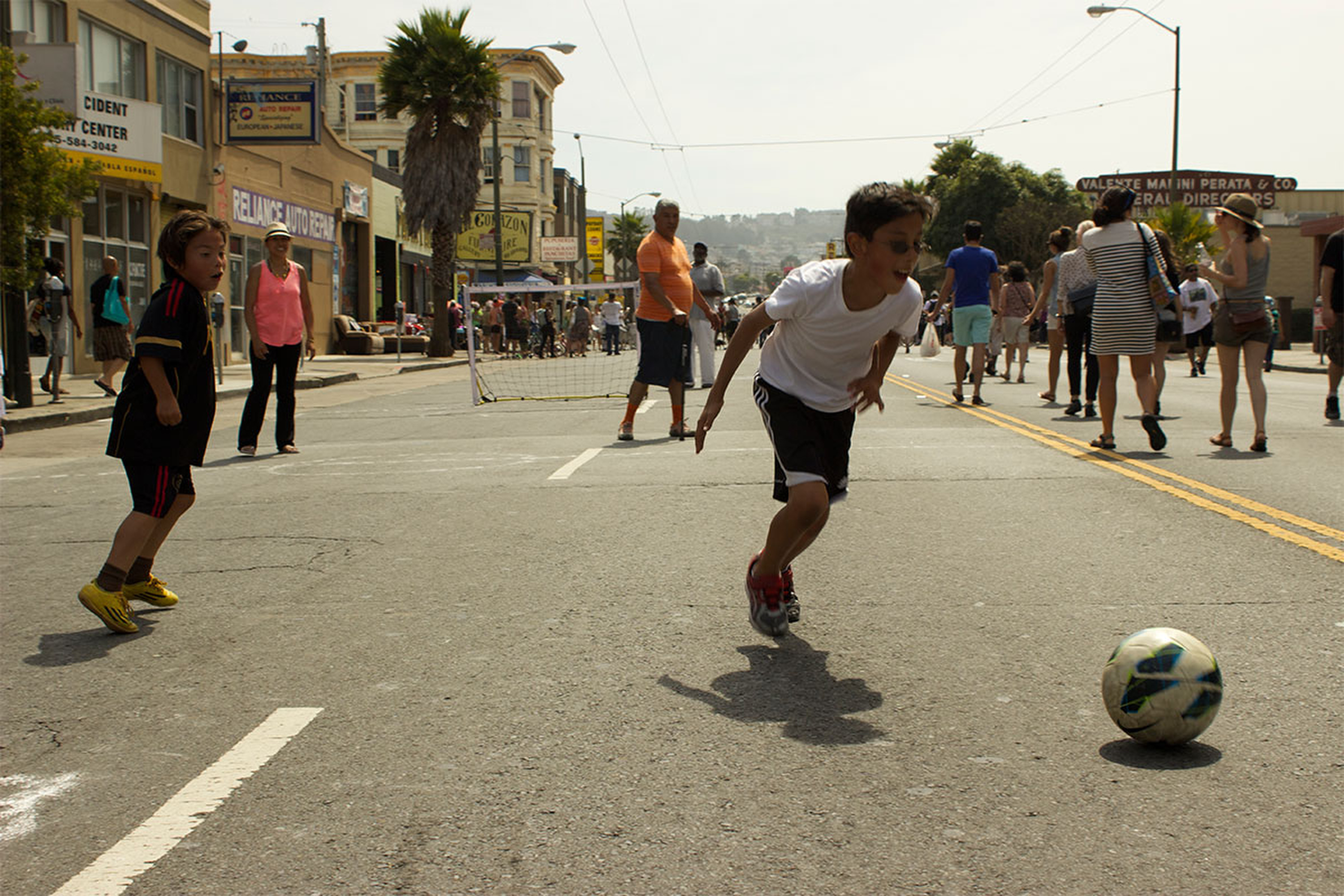 Two kids play street soccer, with others walking nearby. A boy in a white shirt chases the ball, while a boy in a black jersey watches. There's a mini goal ahead.